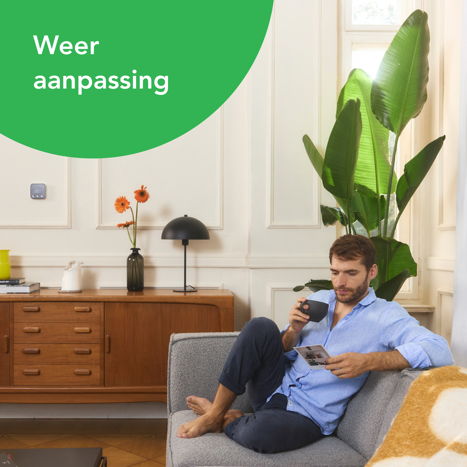 Man op de bank, koffie drinkend, naar een foto kijkend, met een slimme thermostaat aan de muur.