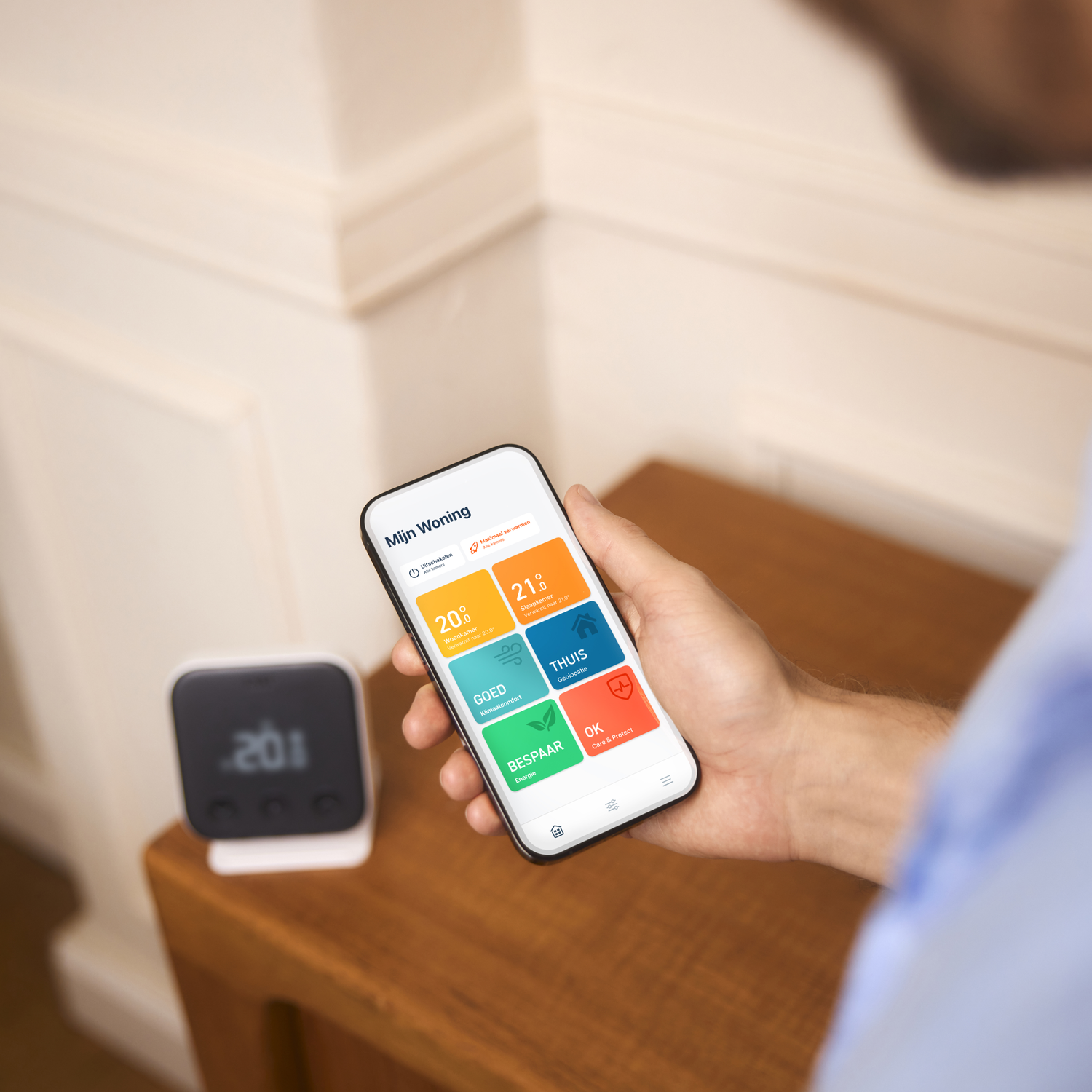 A person holding a smartphone displaying home automation app. A smart thermostat is also on the table.