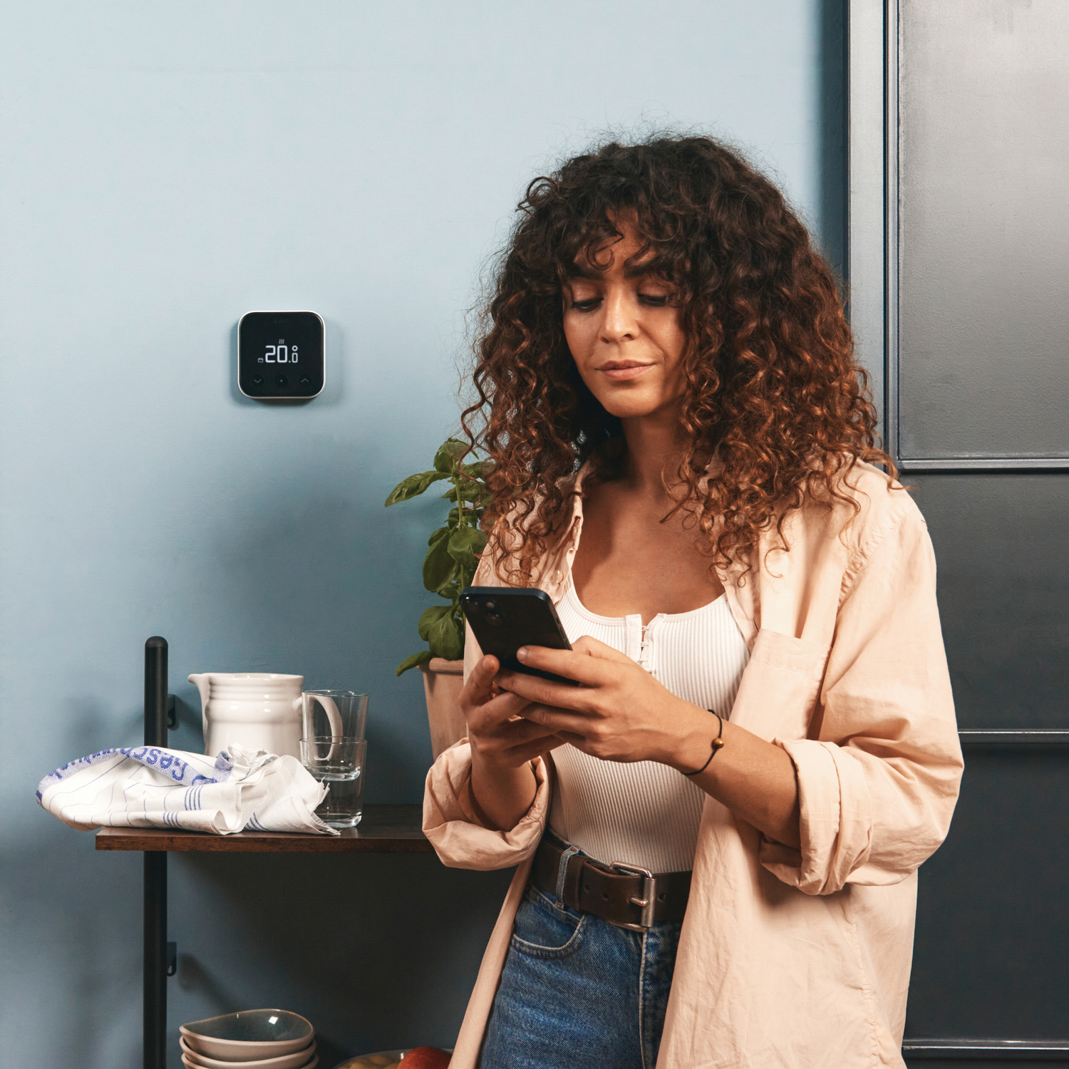 Woman in a light pink shirt using a phone in front of a smart thermostat on a blue wall.