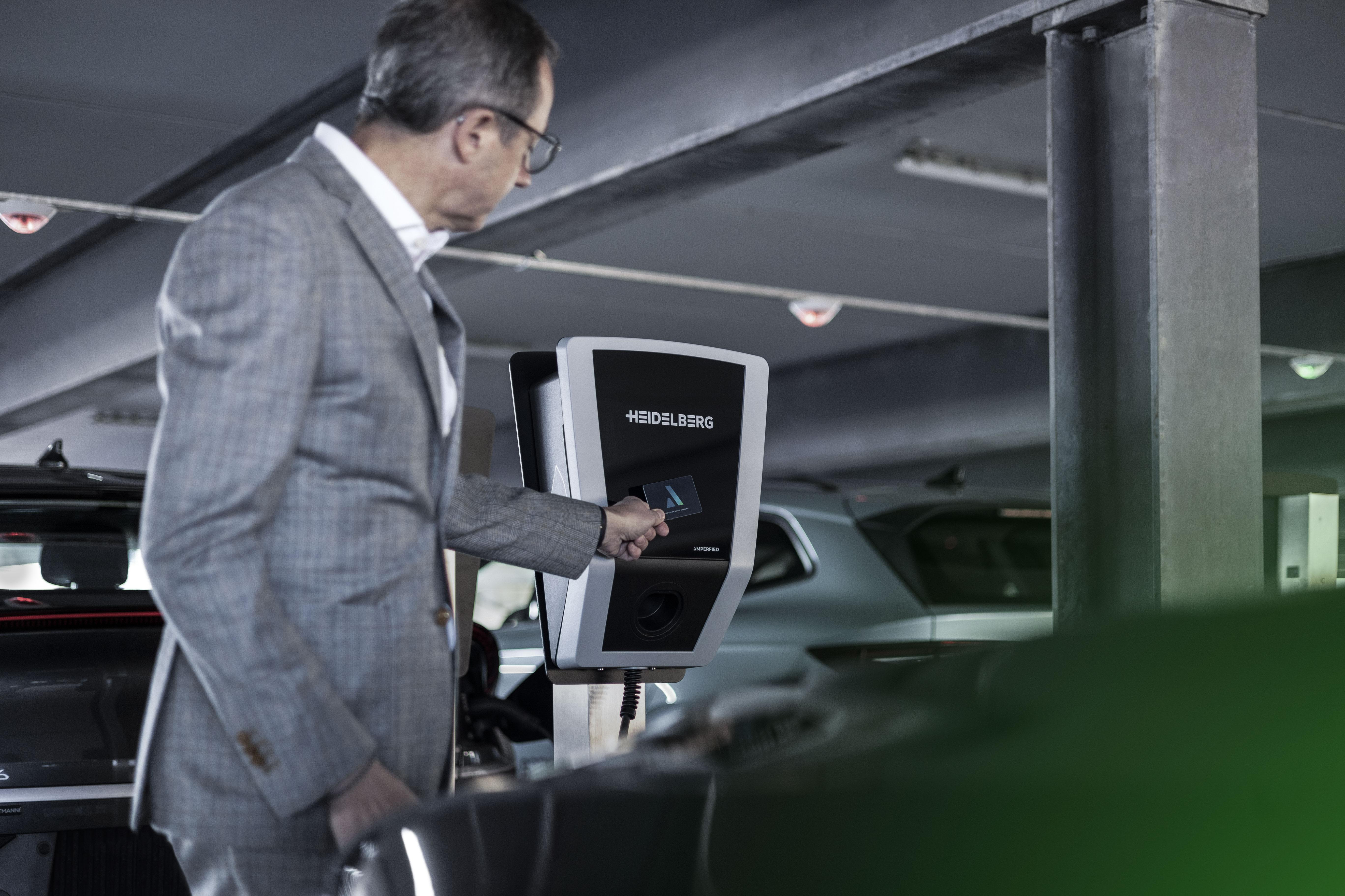 Ein Mann im Anzug benutzt eine Karte an einer Ladestation in einer Parkgarage, Autos sind sichtbar.