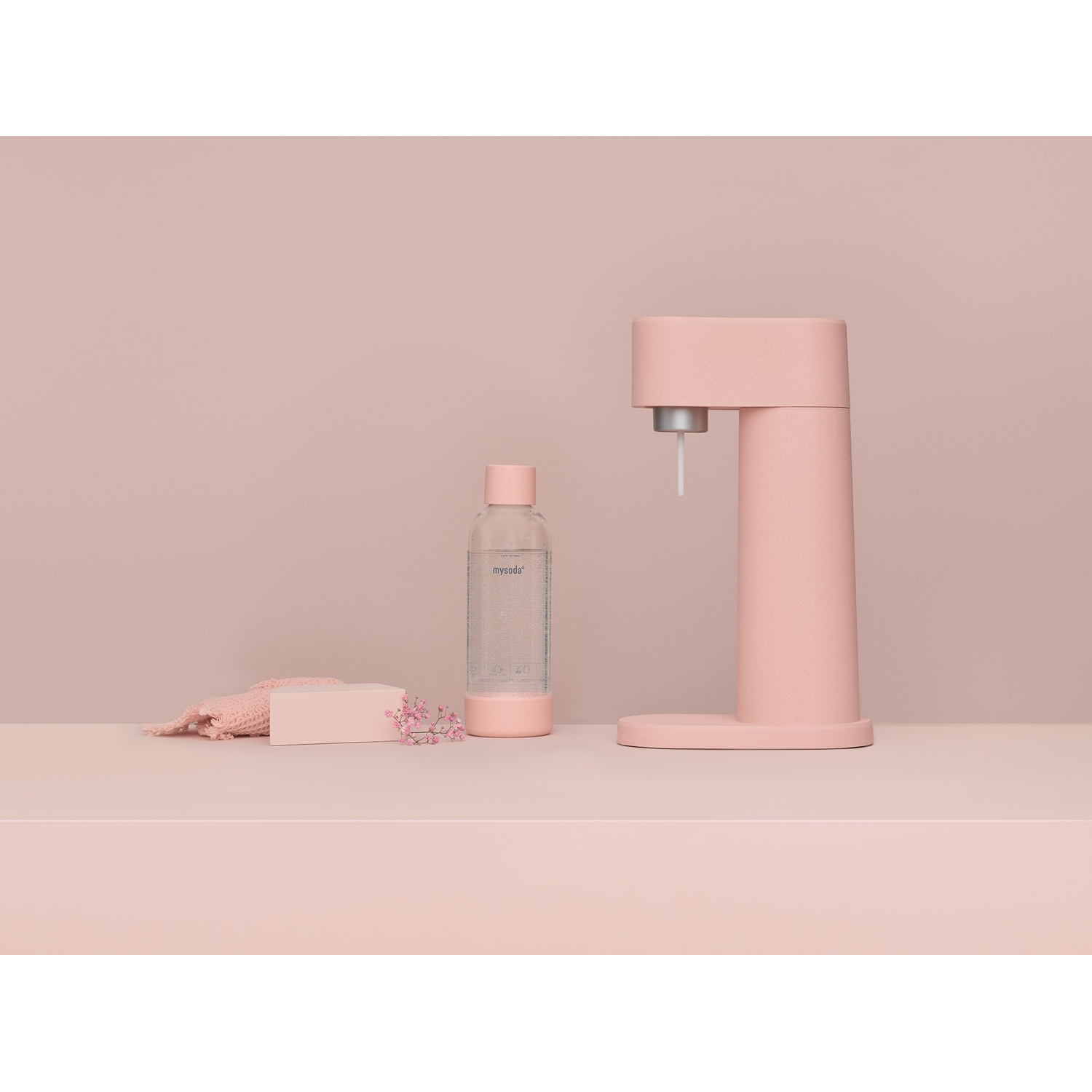 A pink soda maker, water bottle, and a soap bar on a pink surface, against a pink background.