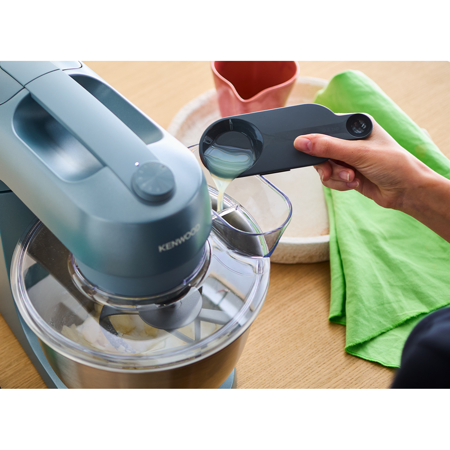 Person pouring liquid into a Kenwood mixer. Mixer is light blue. There is a spoon and a green cloth.