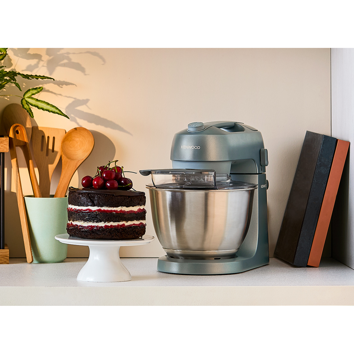 A gray stand mixer next to a cake on a stand. The scene includes wood spoons and a cutting board.