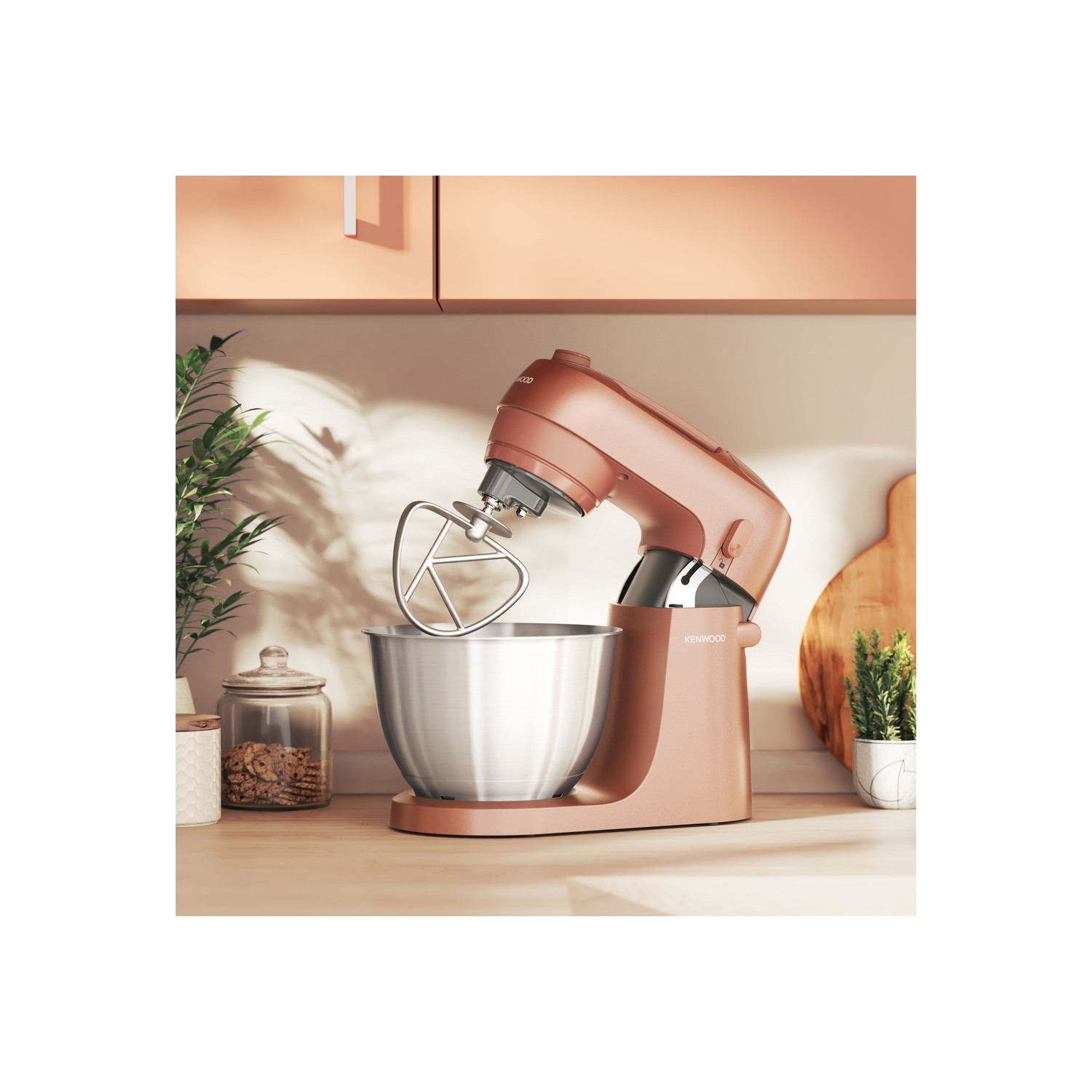 A mixer in a kitchen, in a rose-gold color with a stainless steel bowl, near a jar and cutting board.