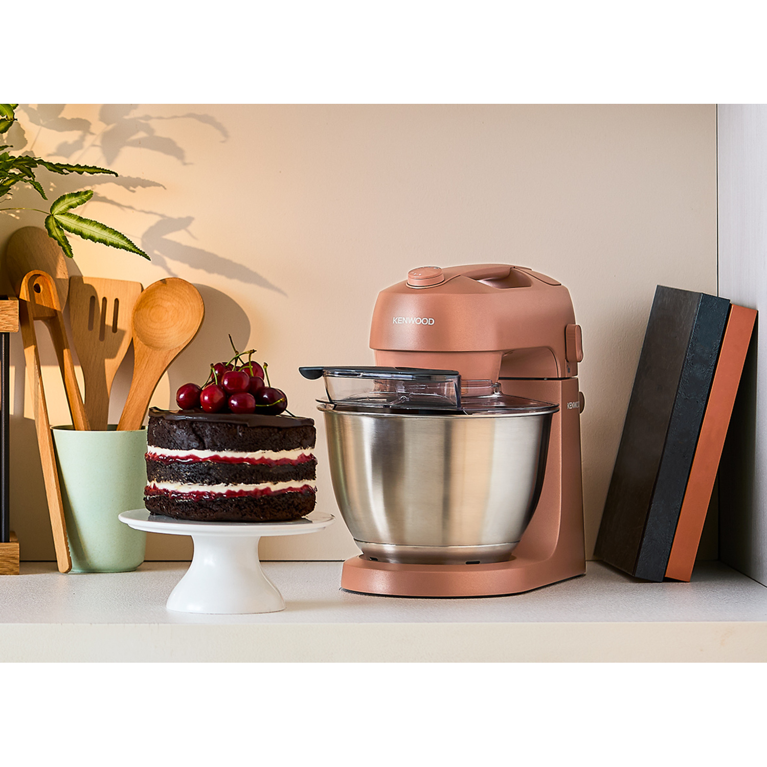 A cake on a white stand, a blender, a wooden spoon, and a plant are on a white surface.