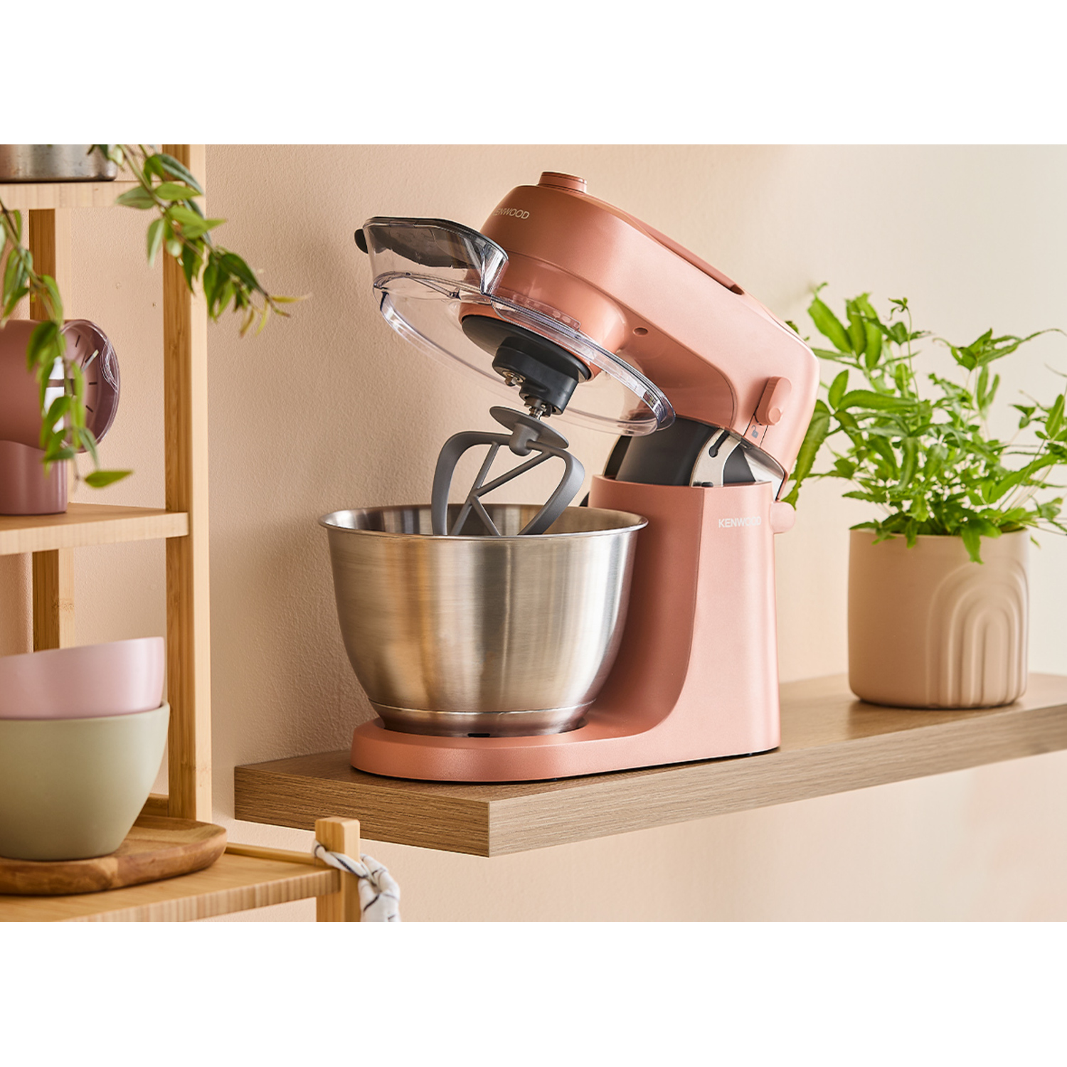 Pink stand mixer on shelf. Silver bowl inside. Plants in background. Kitchen setting.