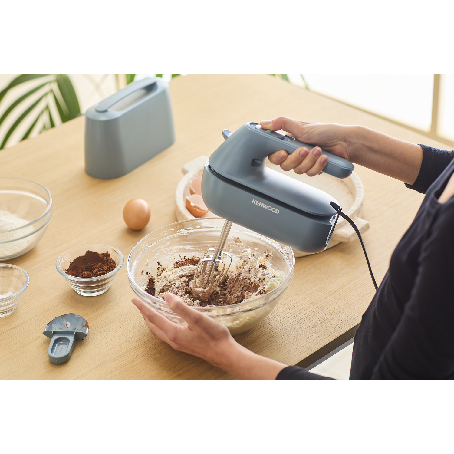 Een persoon die een handmixer gebruikt om ingrediënten in een kom te mengen. De scène bevat bloem en eieren.