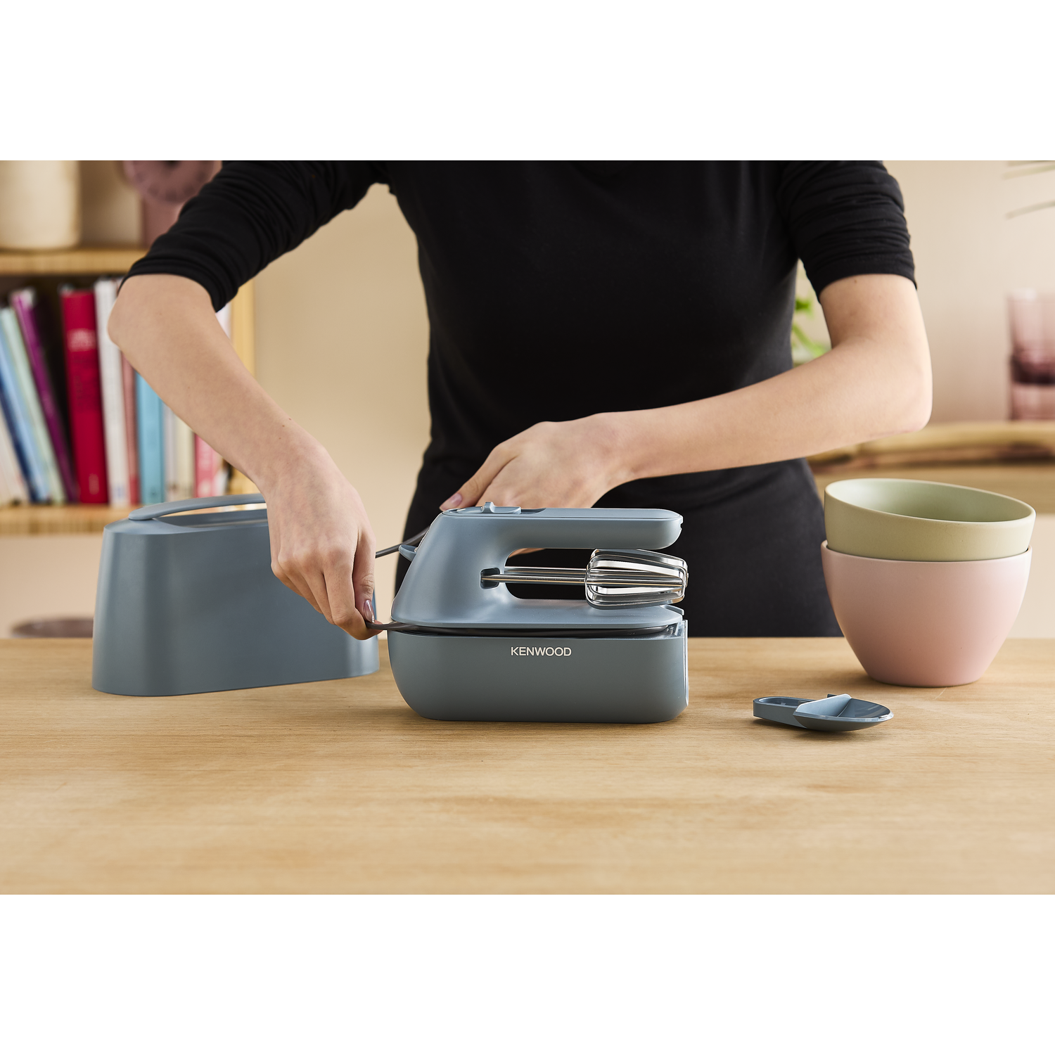Persoon gebruikt een blauwe handmixer op een houten tafel met kommen.