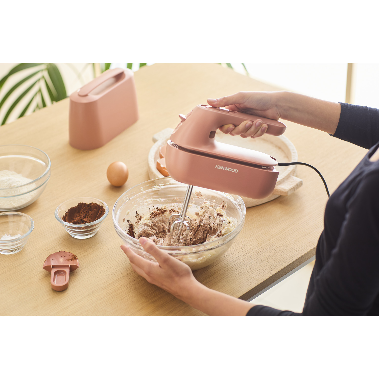 A person is mixing in a bowl with a pink hand mixer. Ingredients on the table include eggs and cocoa powder.