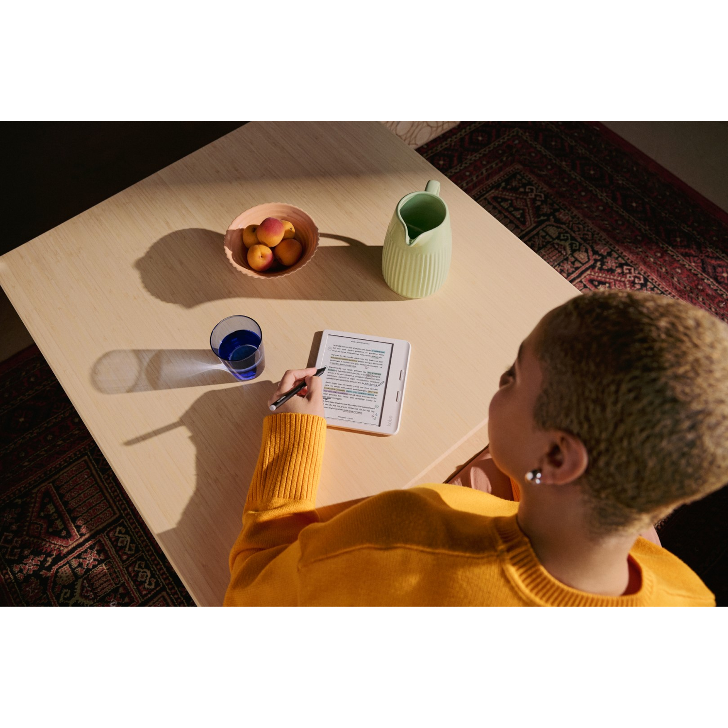 A person is sitting at a table, reading an e-reader, with fruits, a pitcher, and a glass.