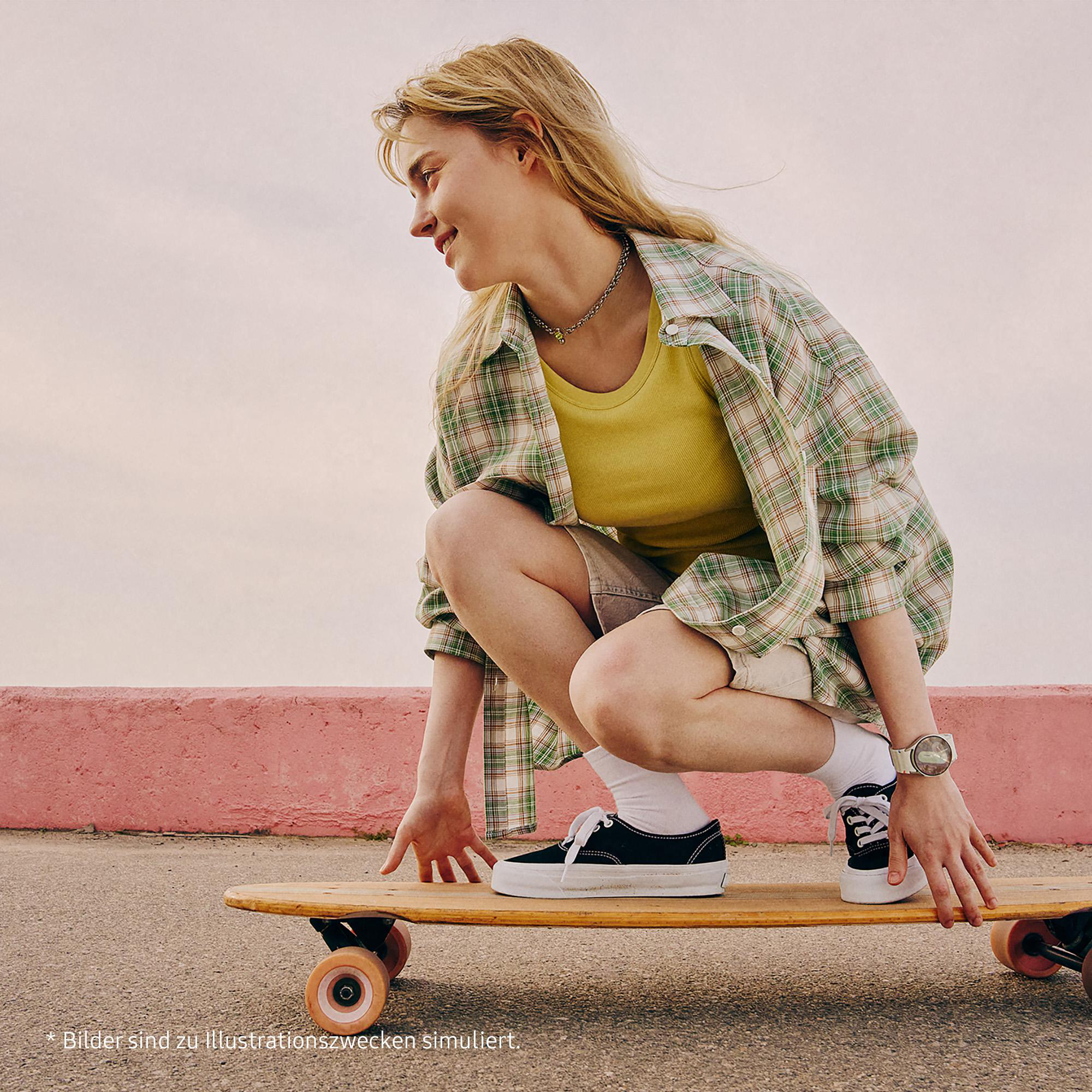 Frau hockt auf einem Skateboard. Sie trägt ein kariertes Hemd, ein gelbes Top, Shorts und schwarze Schuhe. Pinke Wand.