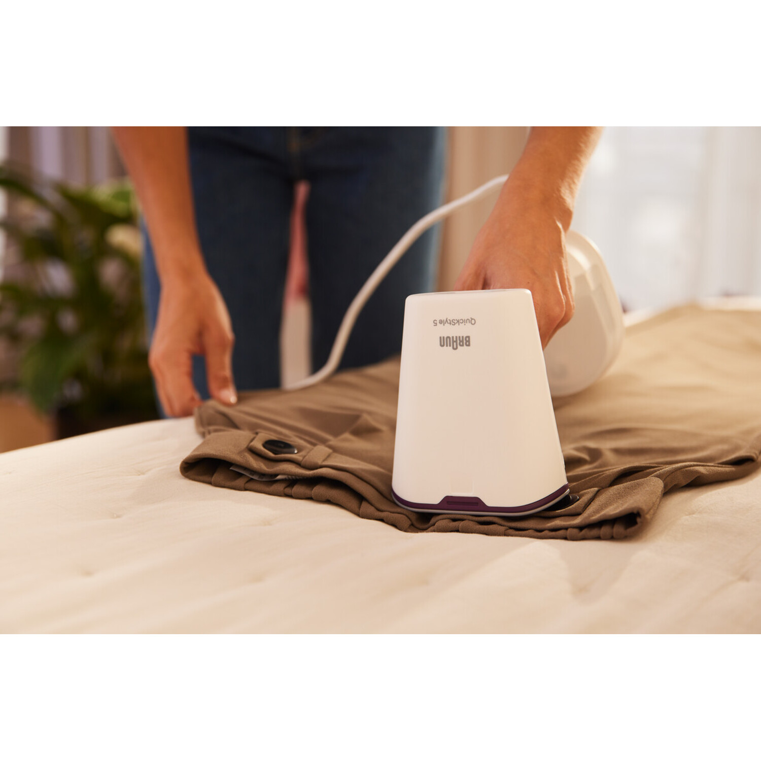 Une personne repassant un pantalon marron sur un lit beige. Un fer à repasser blanc est à droite.