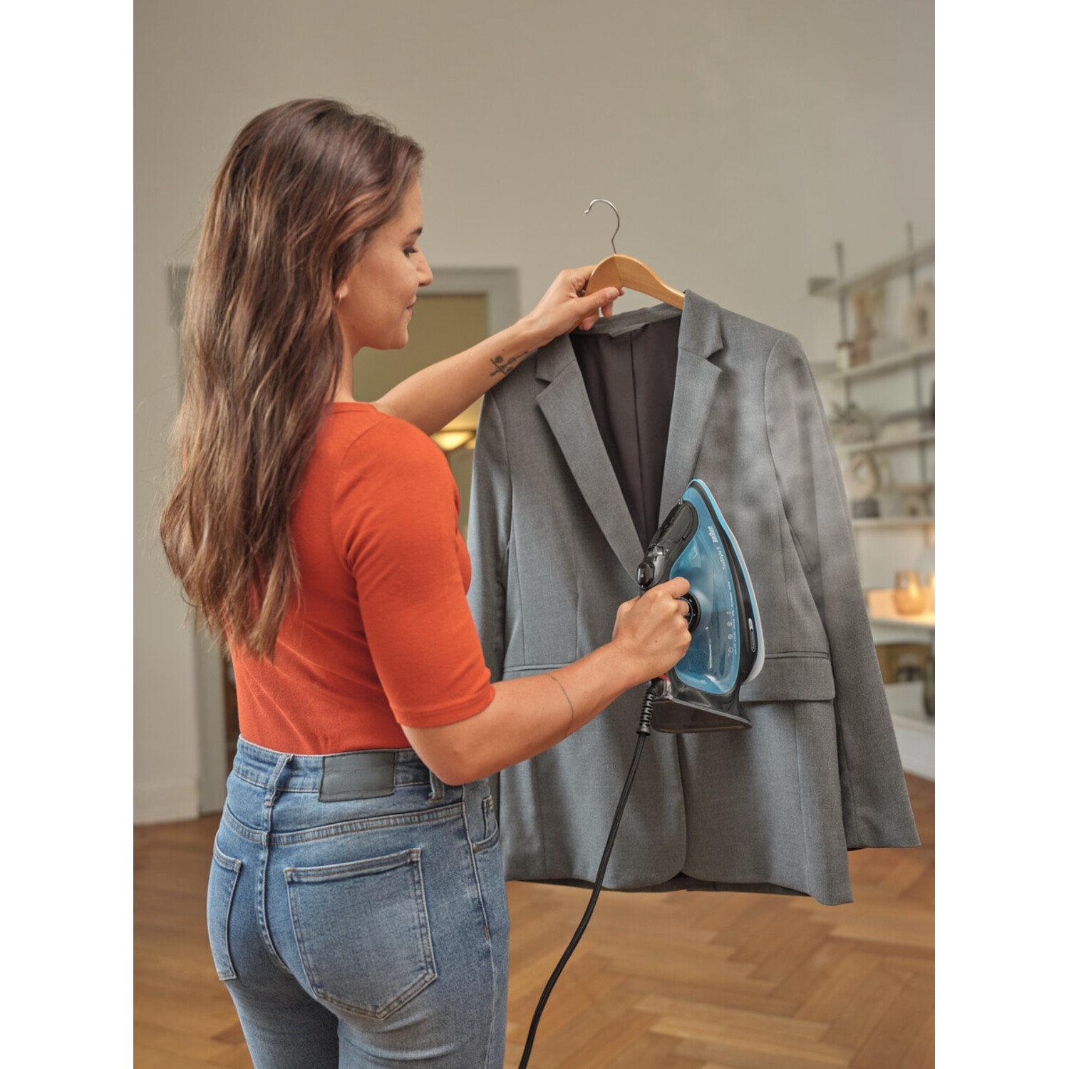 Vrouw die een grijze jas strijkt met een blauwe strijkijzer. Kleding op een hanger.