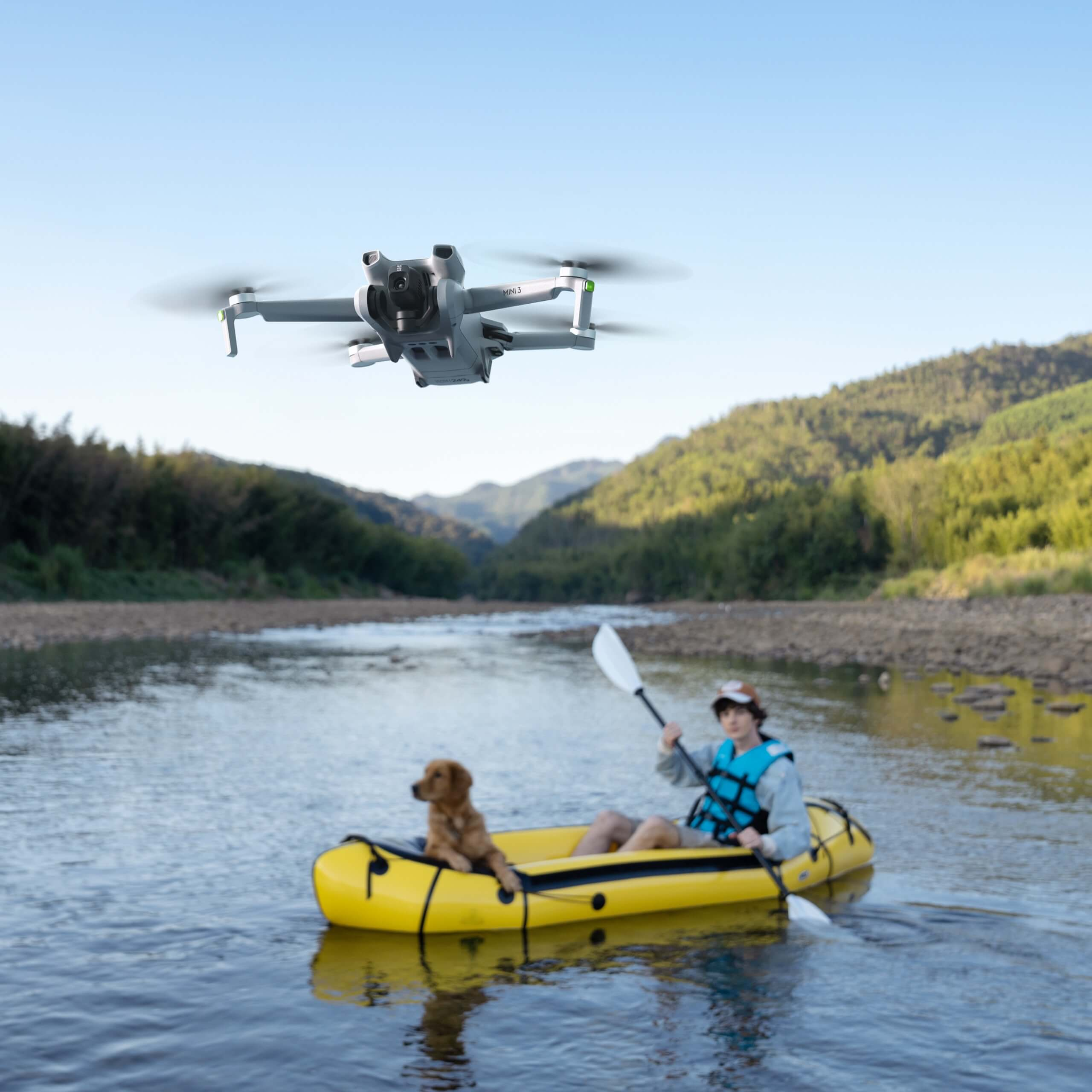 Eine Drohne fliegt über einem Fluss mit einem Kajakfahrer, Hund und Bergen im Hintergrund.