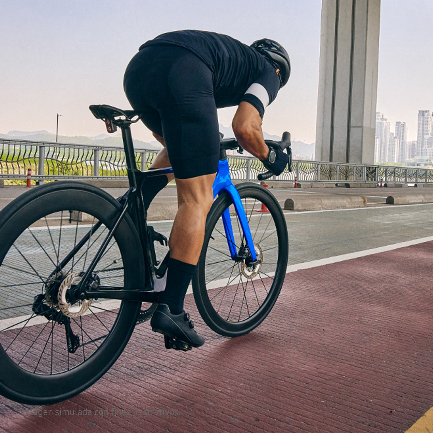 Ciclista con equipo negro en bici azul, sobre carretera roja bajo puente, ciudad al fondo.