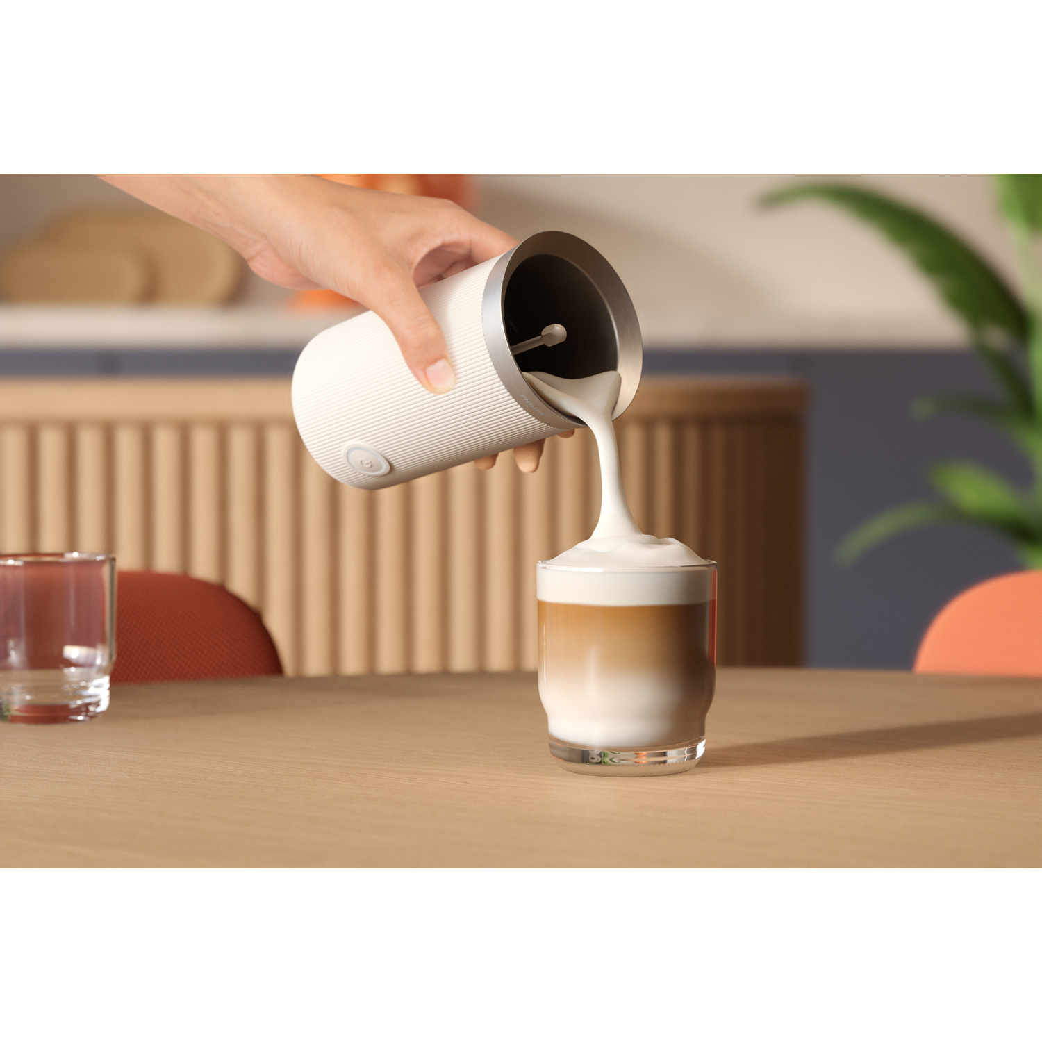 Een persoon schenkt melkschuim in een gelaagde koffie, op een houten tafel, met een glas ernaast.