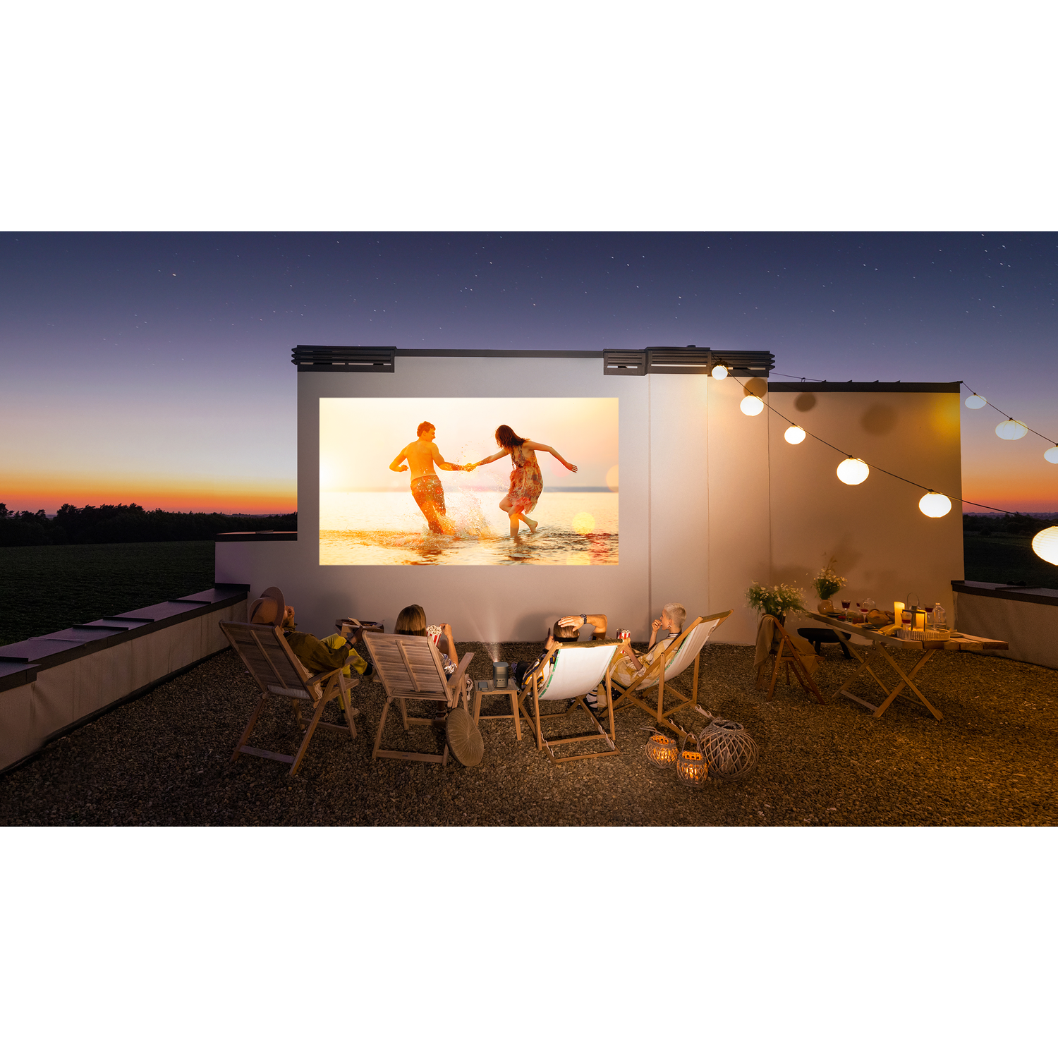 People watch a movie on a projector screen outdoors at night. String lights are on.