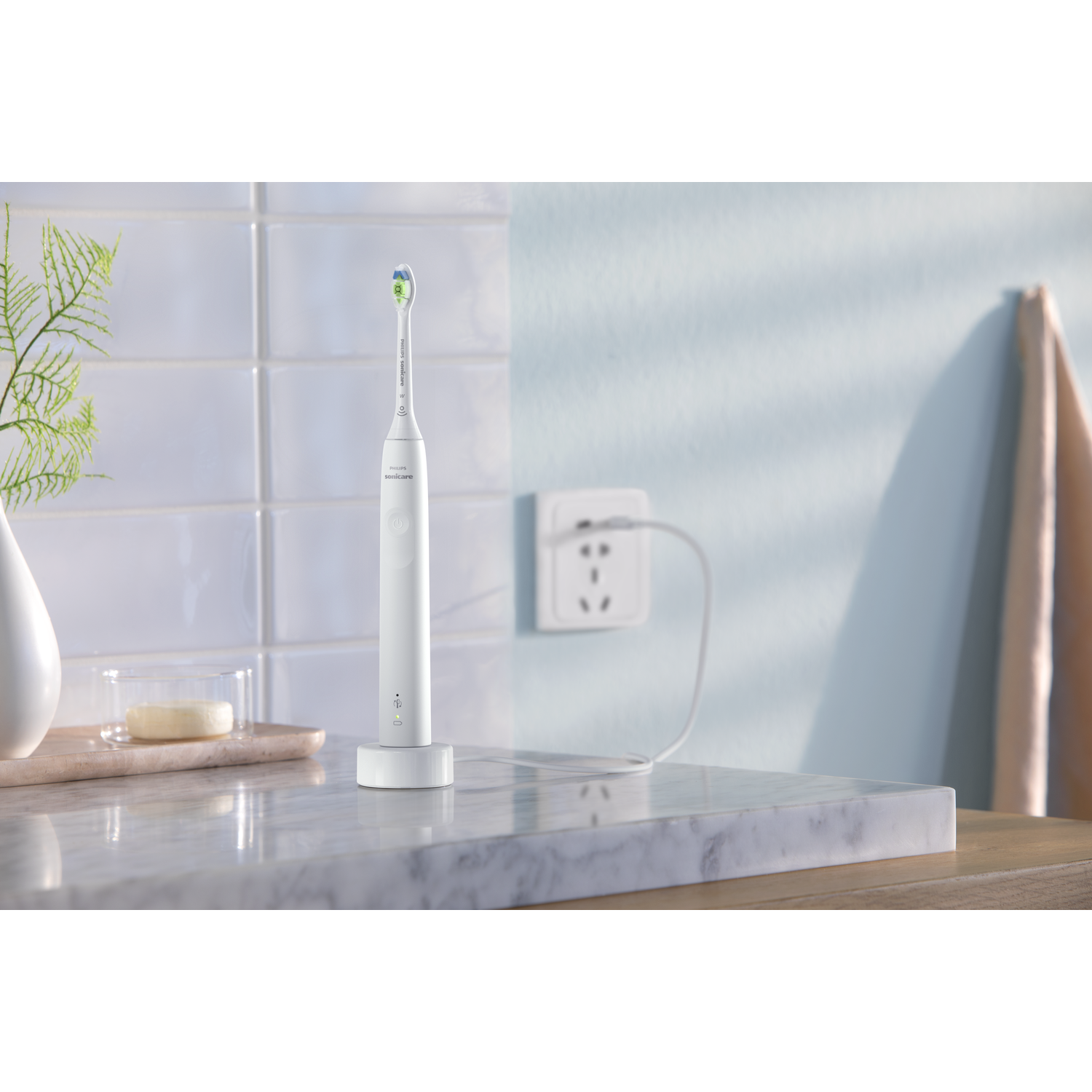 White electric toothbrush on a charger, with a glass and a plant in the background.