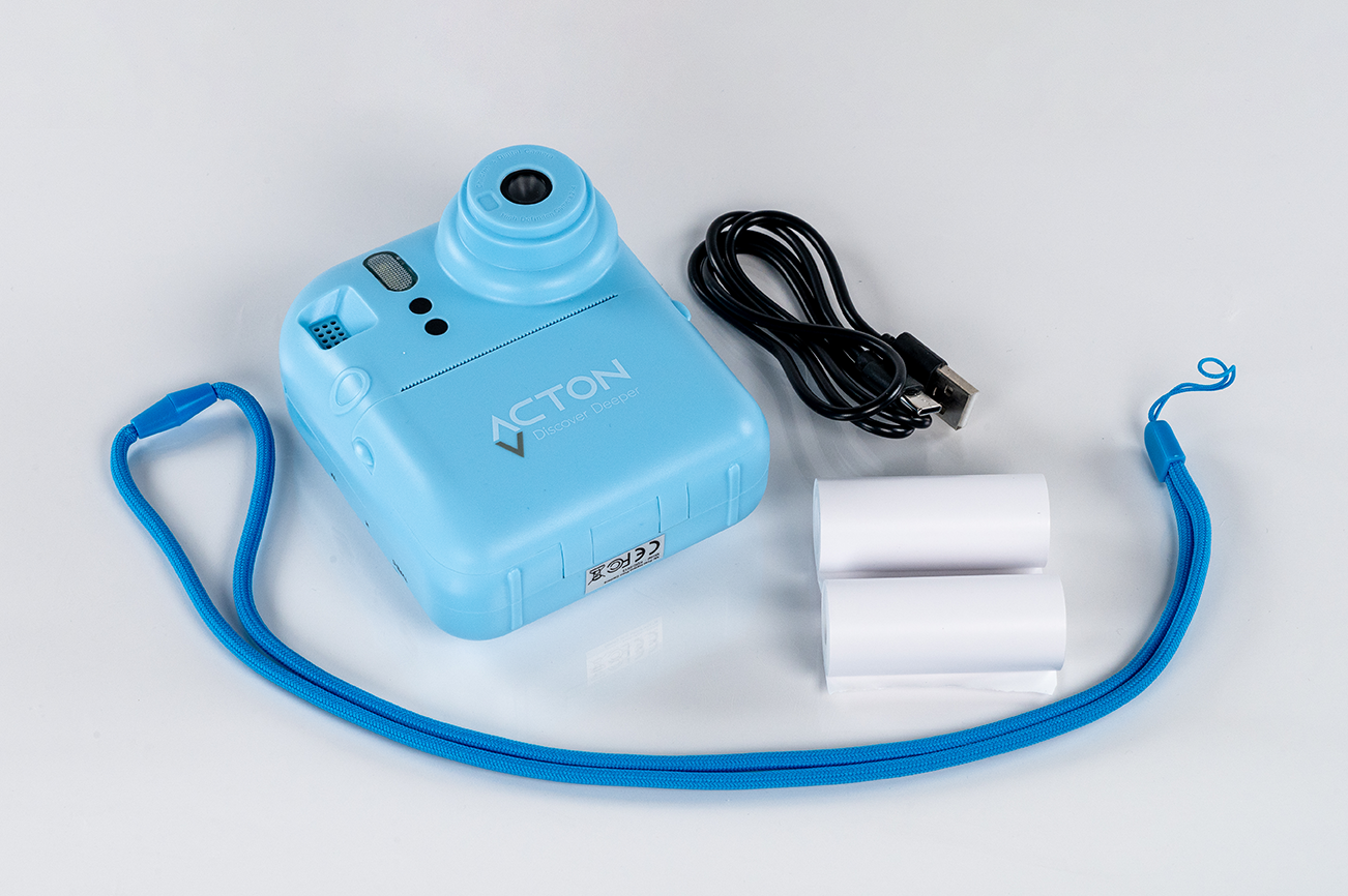 Mavi anında fotoğraf makinesi, aksesuarlarla: USB kablosu, bilek kayışı ve iki rulo kağıt.