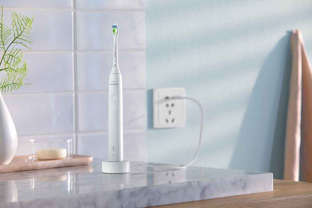 An electric toothbrush on a charging base, next to a wall with a power outlet and a towel.