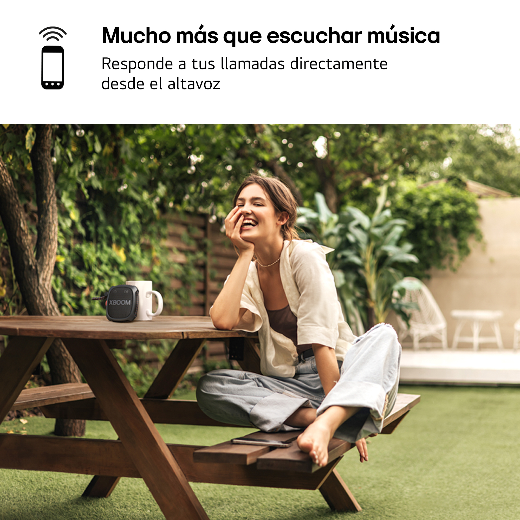 Una mujer sonriendo sentada en una mesa de madera al aire libre. Un altavoz y una taza están sobre la mesa.