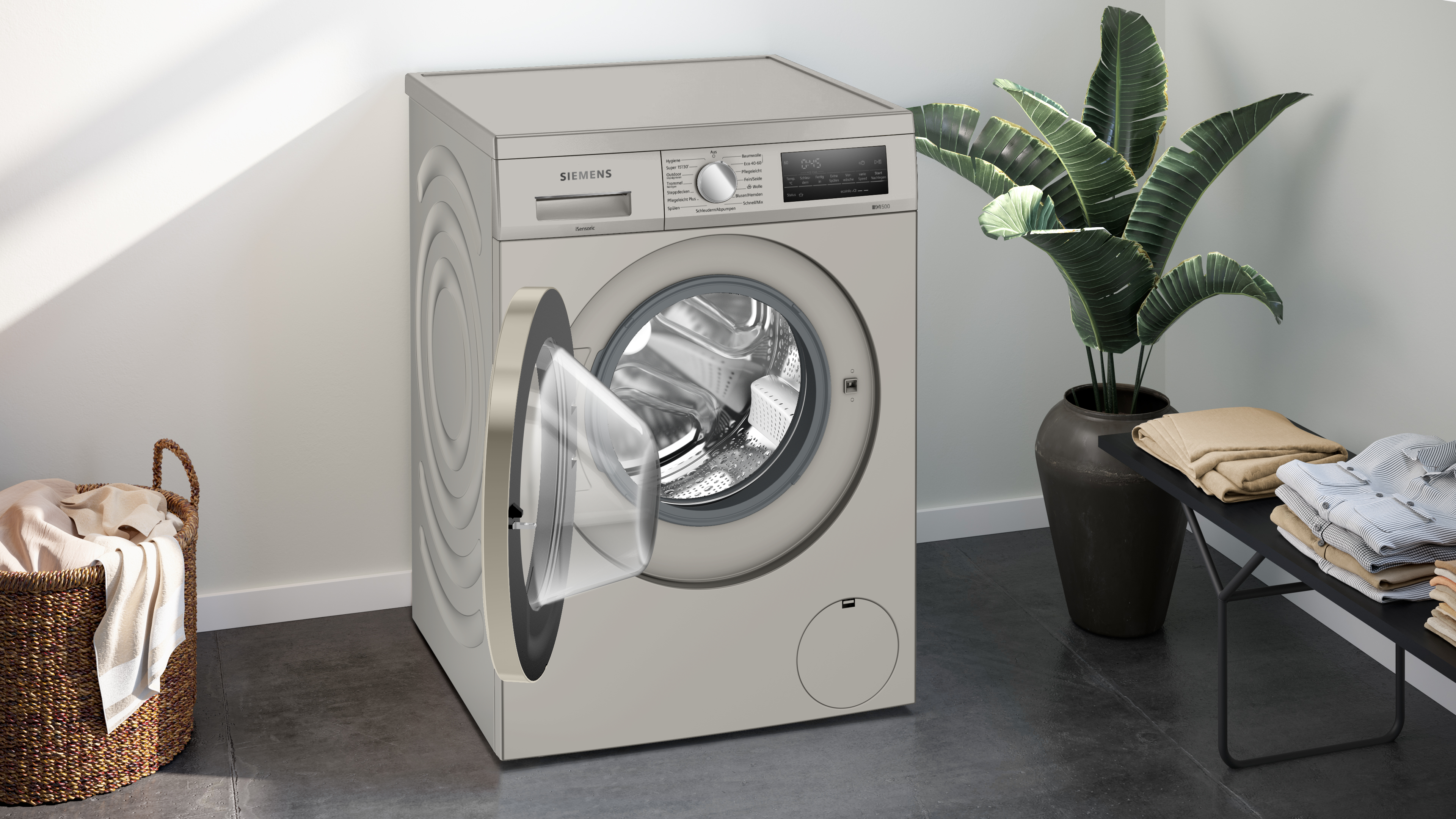 Eine Waschmaschine mit offener Tür. Es gibt einen Wäschekorb, eine Pflanze und gefaltete Kleidung.