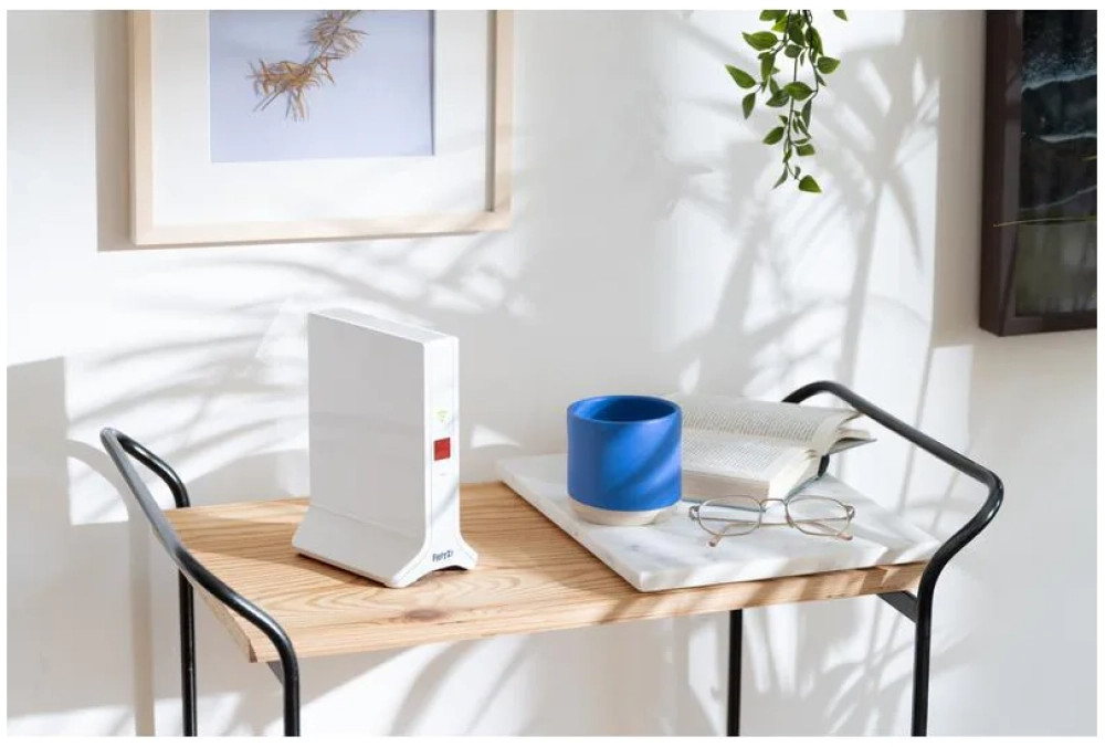 Routeur blanc sur une étagère en bois avec un livre, des lunettes et une tasse bleue. Un tableau est accroché au-dessus.