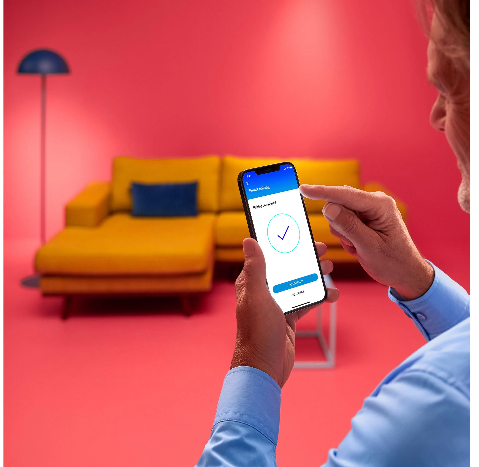 Mann hält ein Telefon, das einen Pairing-abgeschlossen-Bildschirm anzeigt. Gelbes Sofa und rosa Hintergrund.