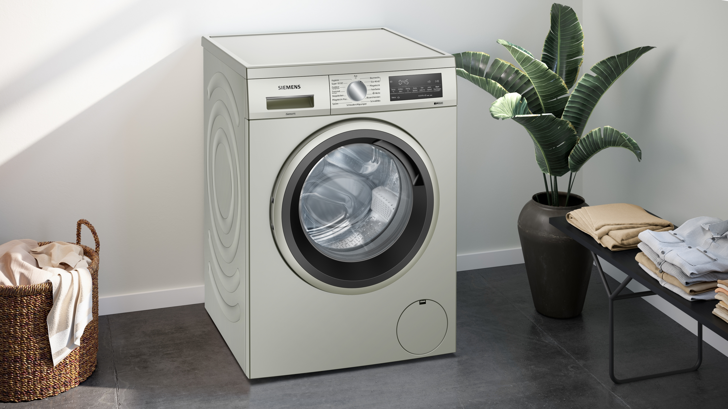 Eine beige Waschmaschine neben einer Pflanze und einem Wäschekorb. Der Raum hat weiße Wände.
