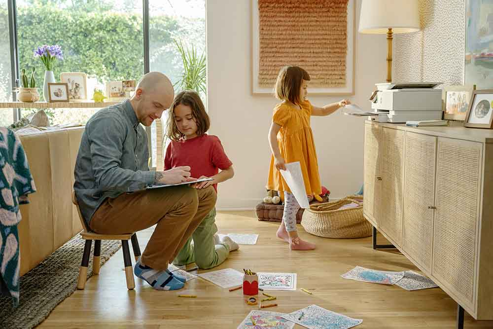A family is in a room. A man sits with a child drawing. Another child is near a printer.