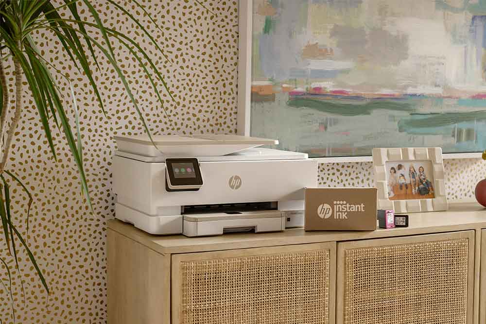 White HP printer on a wooden cabinet. A painting and framed photos are behind it.