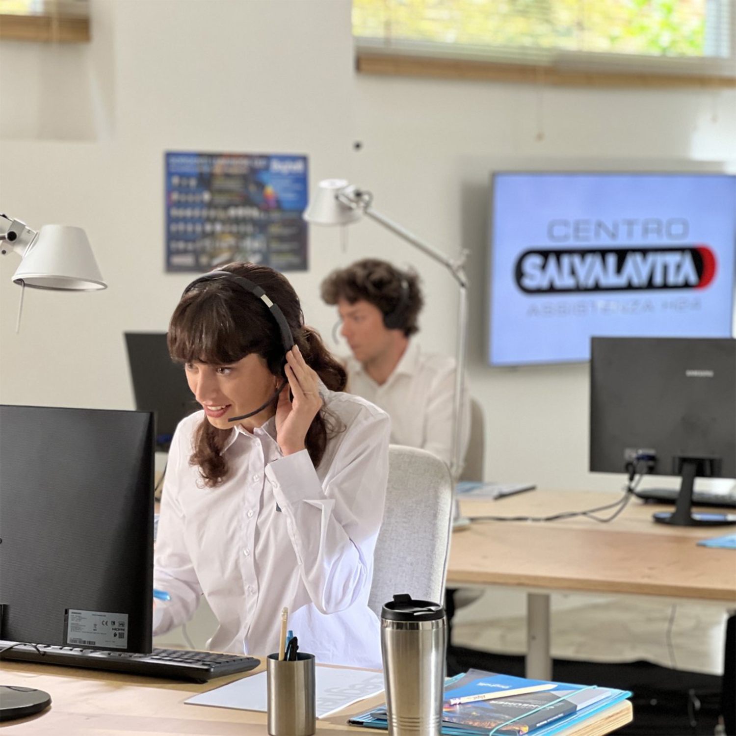 Due persone in un call center, una con le cuffie, che guardano gli schermi dei computer.