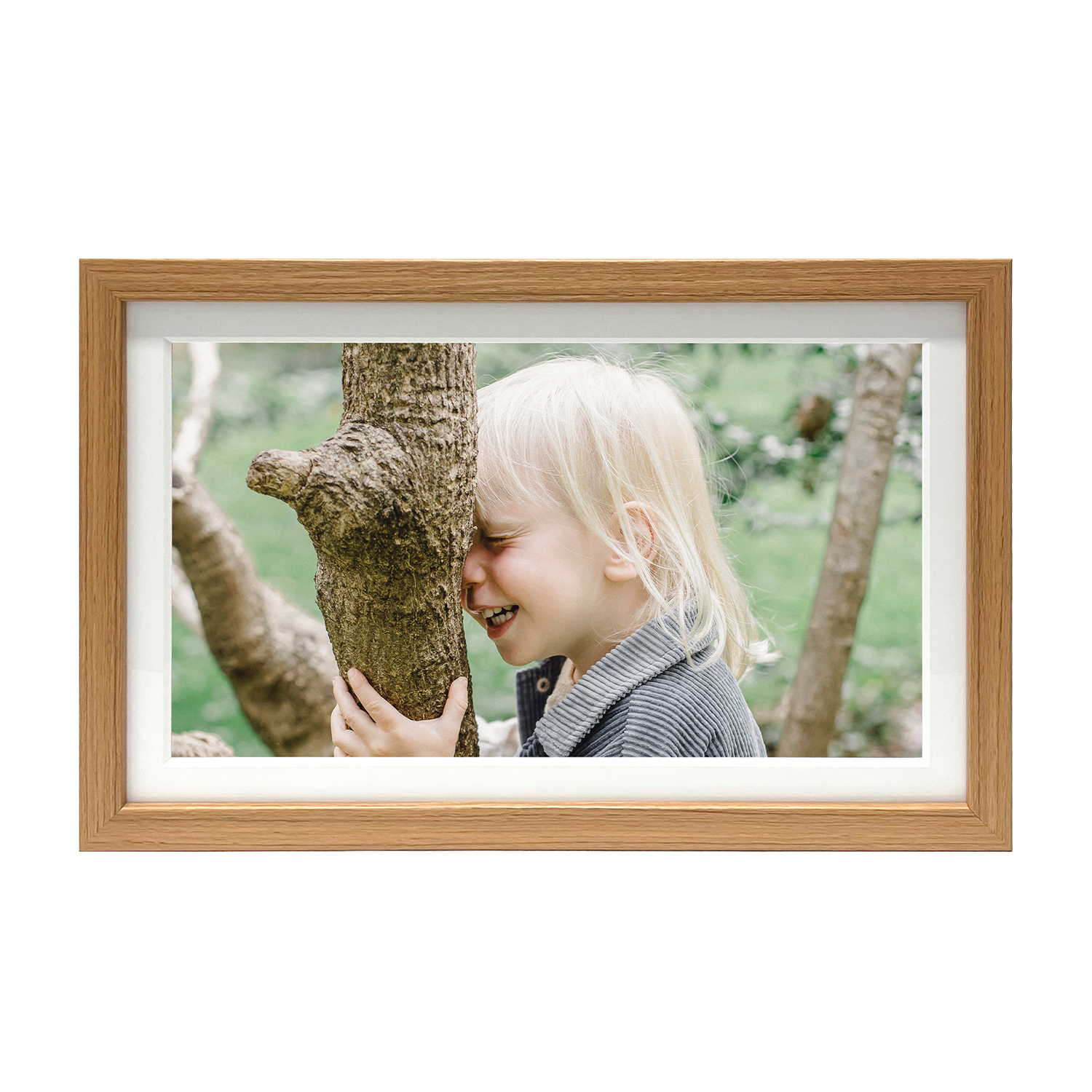 Bambino con capelli biondi che ride appoggiato a un tronco d'albero in una cornice.