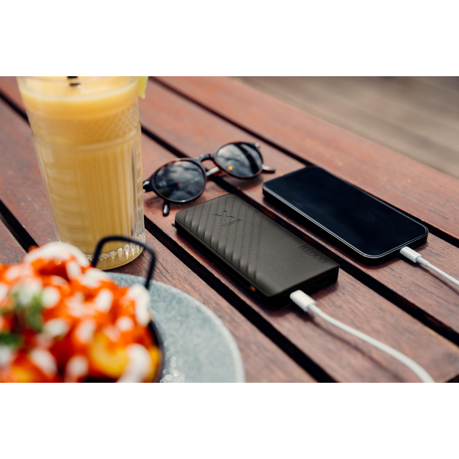 Tabletop scene with a phone, charger, sunglasses, juice, and a snack. The background is blurred.