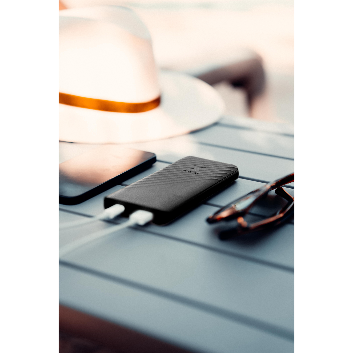 A black power bank with white charging cables, a phone, sunglasses, and a hat.