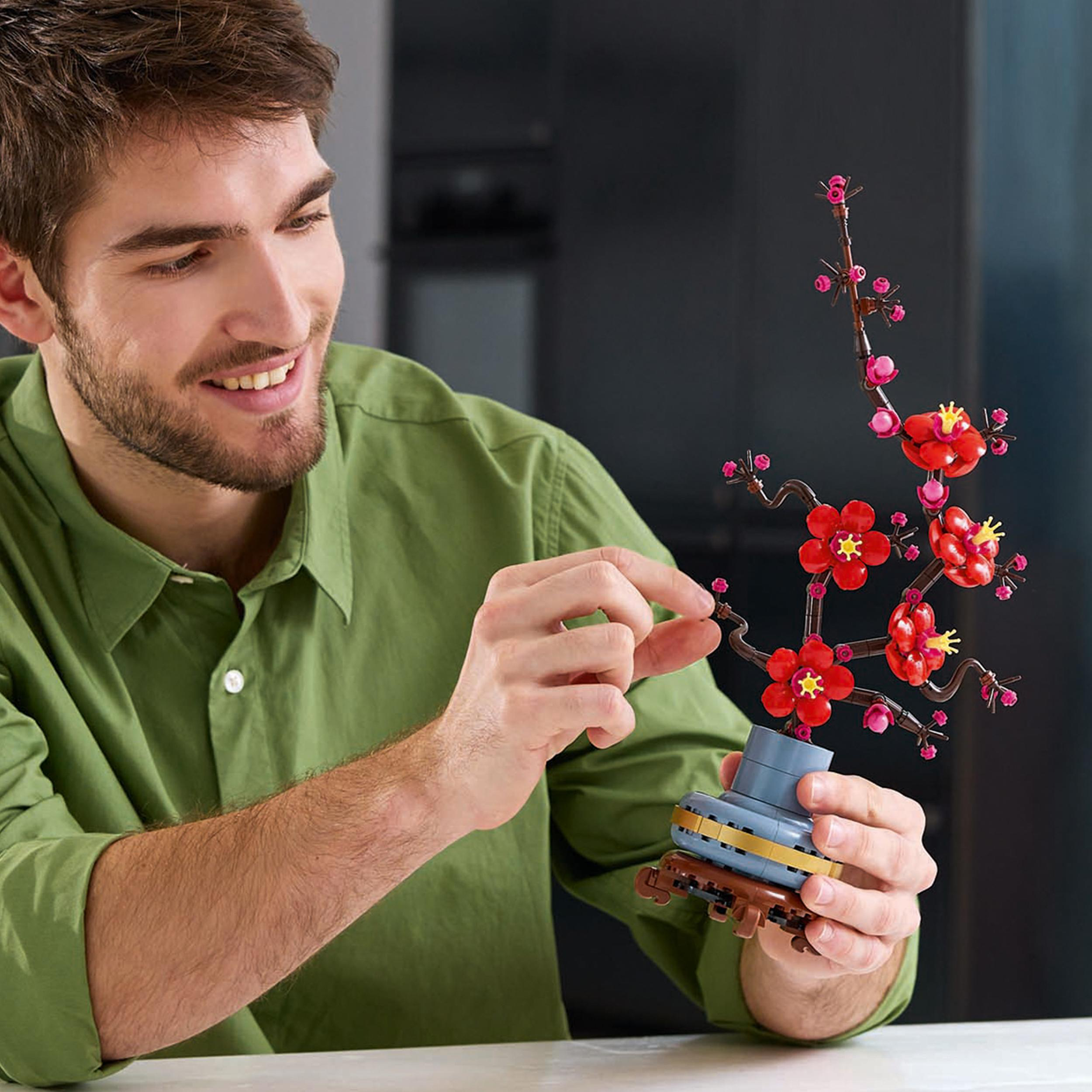 Mann lächelt, baut Lego-Blumen in einer Vase. Rote Blumen, braune Stiele und eine blaue Vase. Weißer Tisch.
