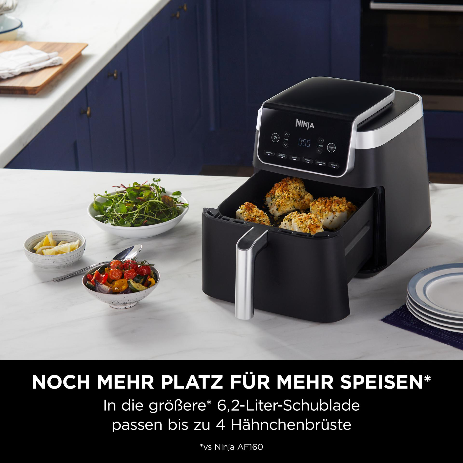 Schwarze Heißluftfritteuse mit Essen und Schalen mit Gemüse und Zitronenscheiben auf einer Theke.