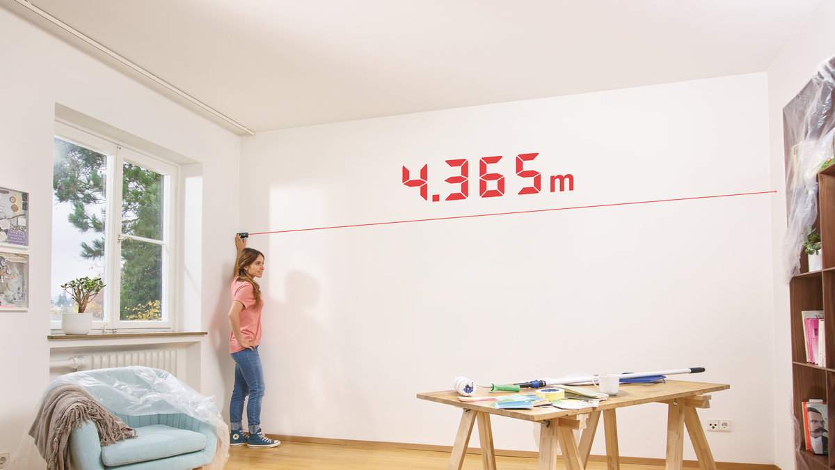 Egy nő egy falat mér lézeres mérőeszközzel. Piros 4.365m számok jelennek meg.