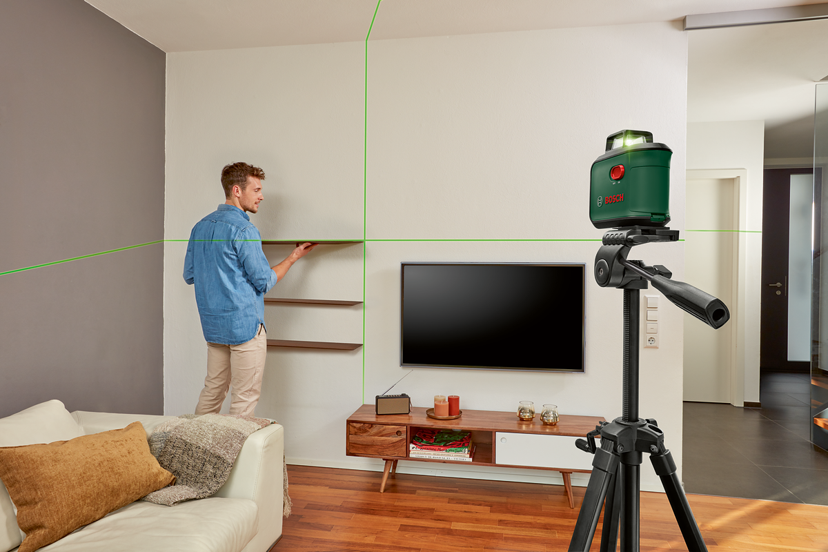 Férfi Bosch szintezővel polcokat szerel. Zöld lézer vonalak láthatók. TV és bútorok láthatók.