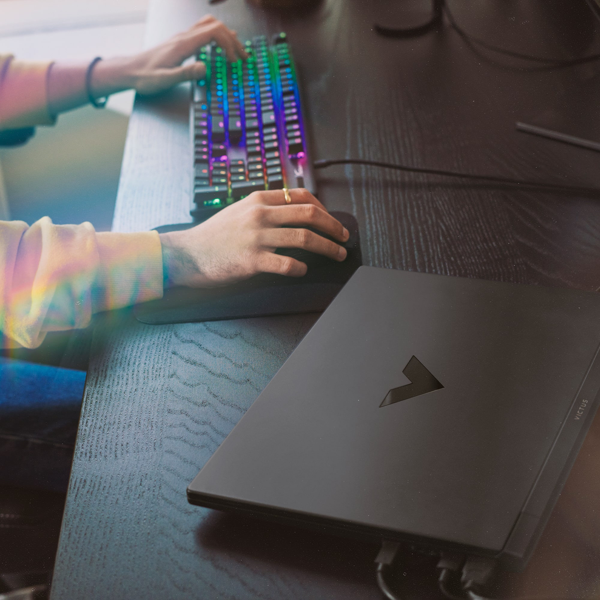 Persona usando un teclado y un ratón, y una computadora portátil. El escritorio es oscuro y el teclado tiene luces coloridas.