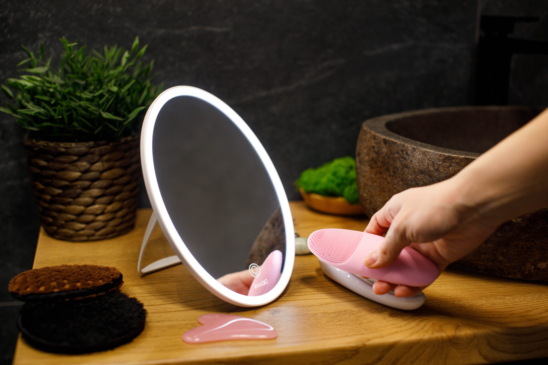 Una mano sostiene un cepillo facial rosa texturizado cerca de un espejo ovalado iluminado sobre una superficie de madera.