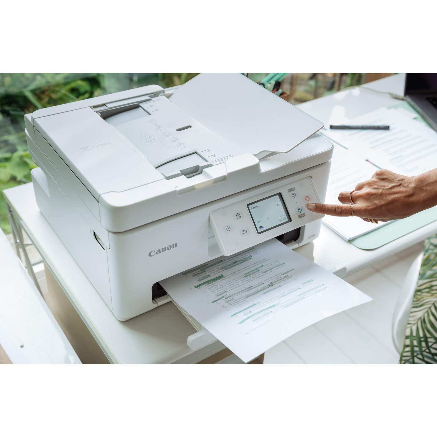 Een witte Canon-printer met de hand van een persoon die op een knop drukt, met bedrukt papier in de uitvoerlade.