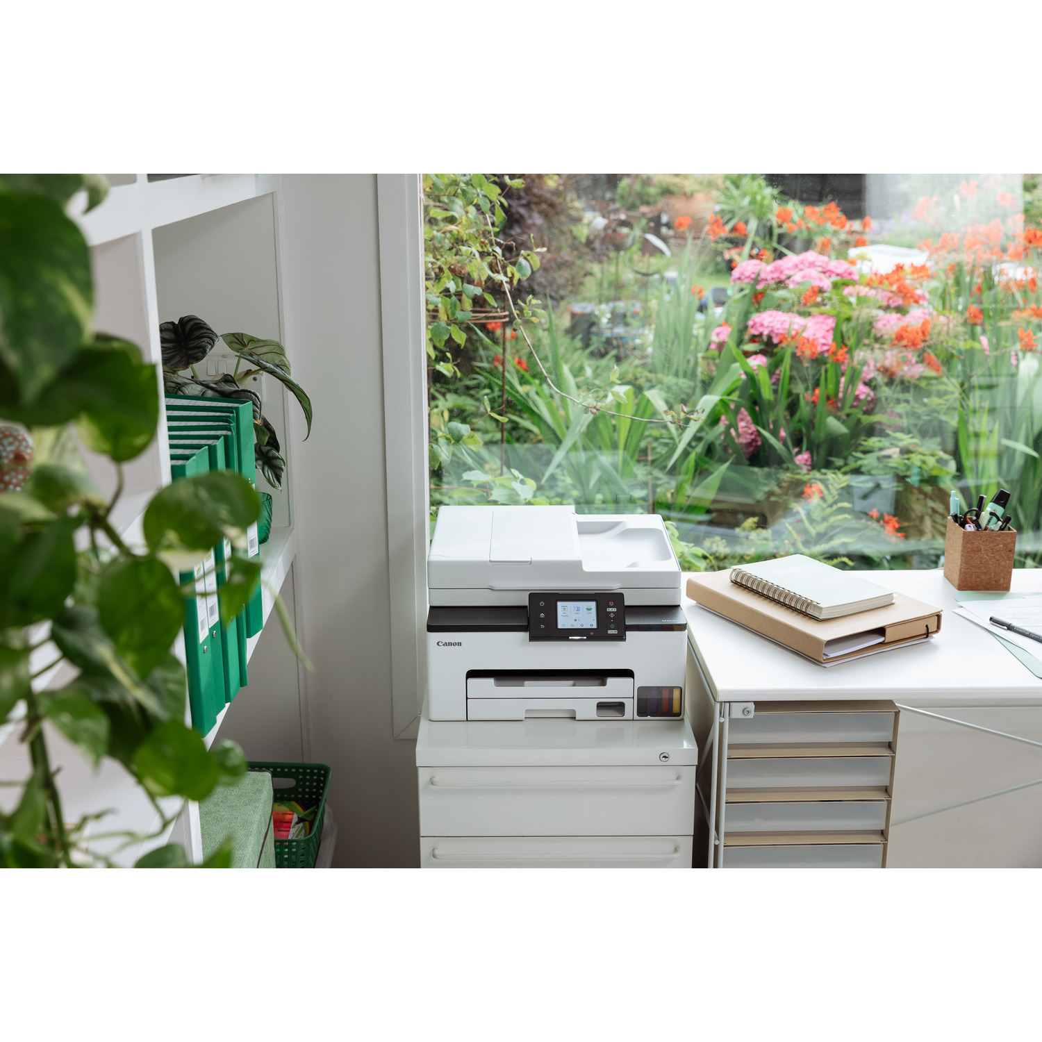 Une imprimante blanche est sur un bureau blanc près d'une fenêtre. Un jardin avec des fleurs est visible.