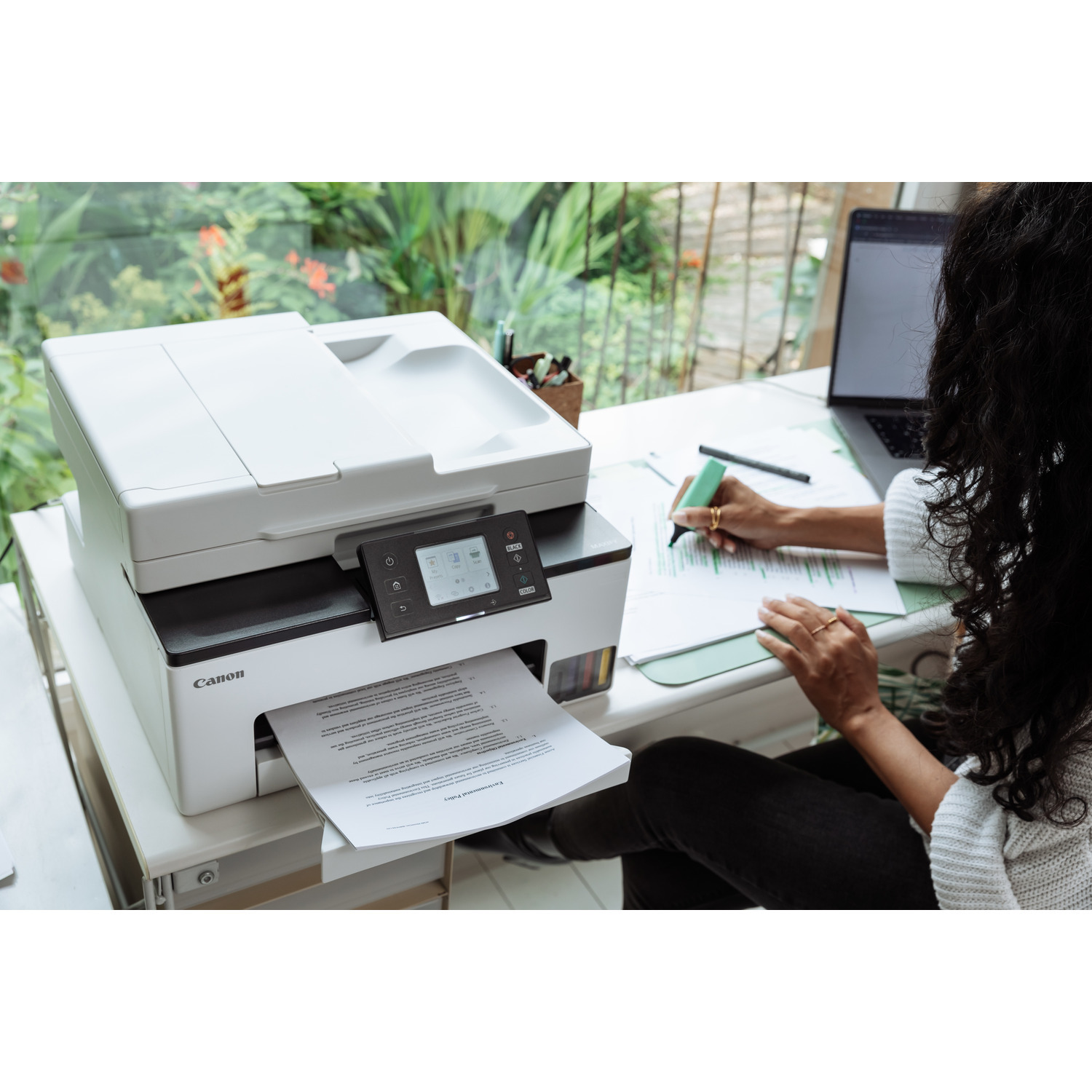 Une femme est assise à un bureau, utilisant une imprimante Canon et écrivant sur un document avec un stylo.