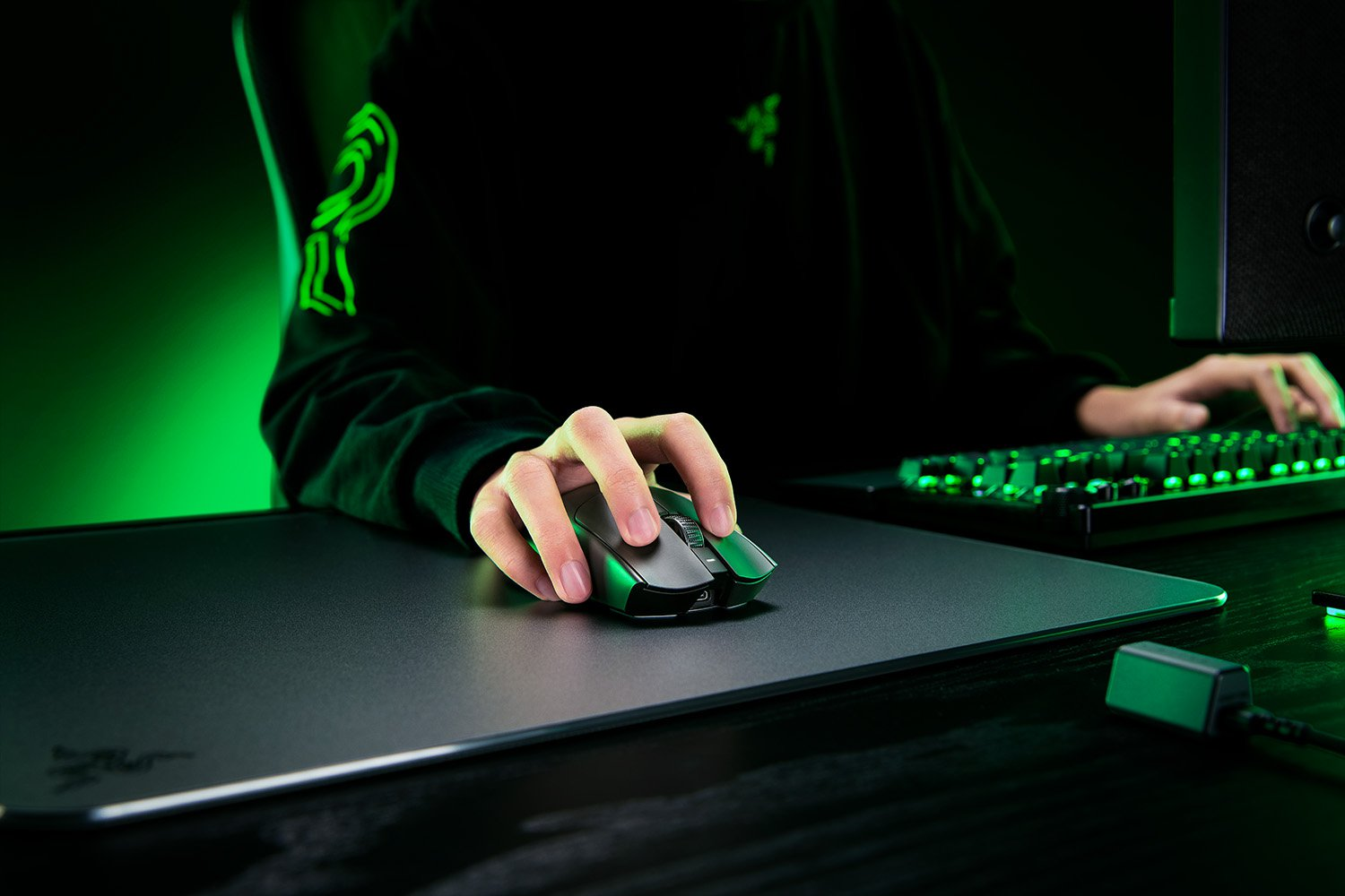 A person's hands using a black Razer gaming mouse on a black mousepad, near a keyboard.