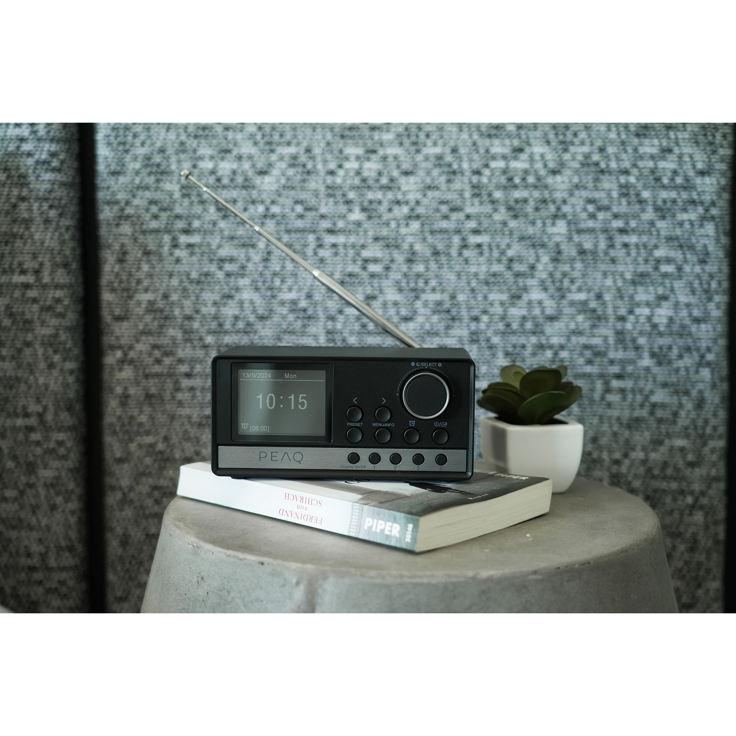 Una radio nera con display digitale, antenna e una piccola pianta su una superficie grigia.