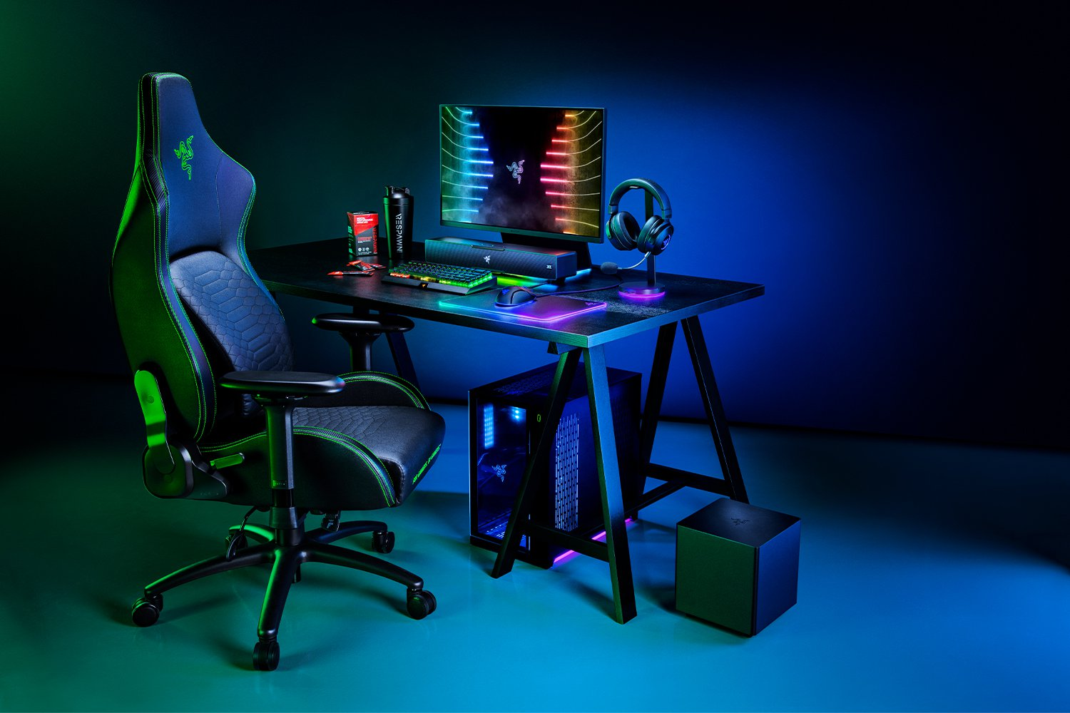 Gaming setup met een zwart bureau, stoel, pc, monitor en accessoires. Blauwe, groene en paarse lichten.