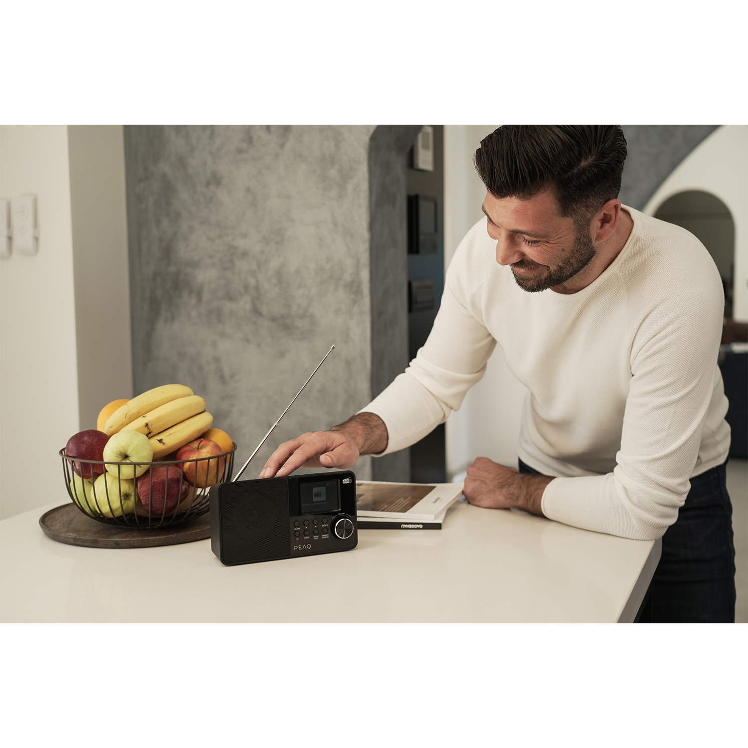 Un uomo sorride mentre interagisce con una radio nera su un piano di lavoro bianco, cesto di frutta in primo piano.