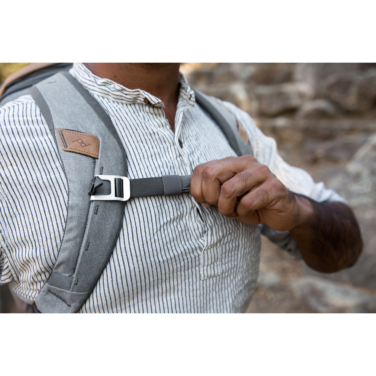 Una persona che regola la tracolla di uno zaino, con uno zaino grigio e una camicia a righe visibili.