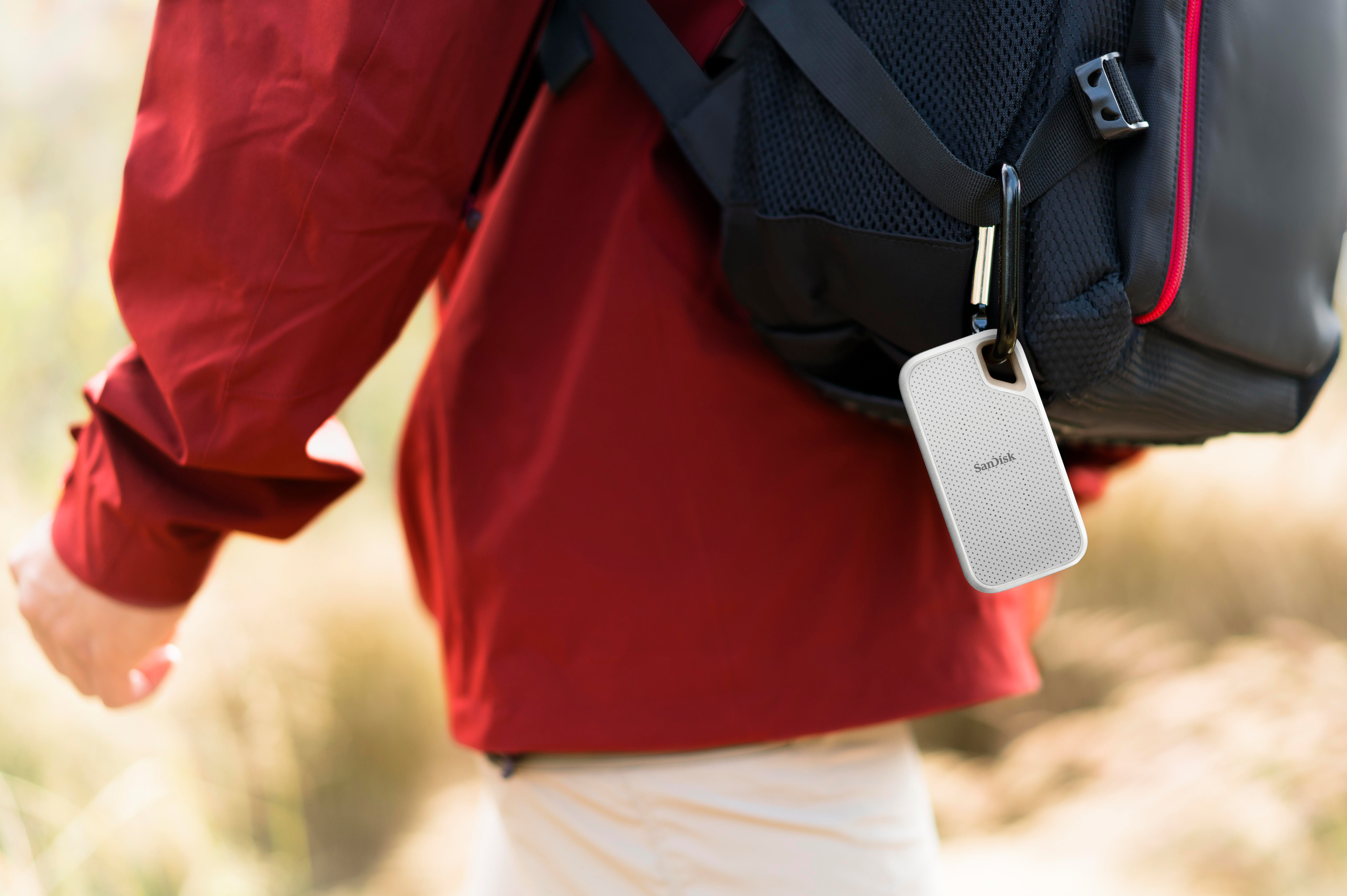 Person in roter Jacke mit schwarzem Rucksack, hält ein weißes und graues Gerät mit Karabiner.